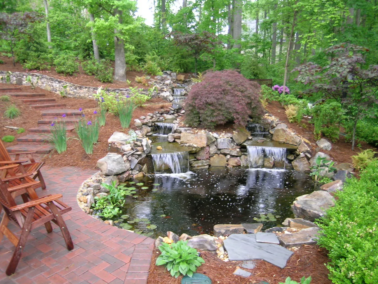 A tranquil garden scene with two wooden chairs, a pond featuring small waterfalls, is surrounded by trees and lush greenery. Thoughtful landscape design incorporates brick and stone paths that gracefully lead through this serene escape. A tranquil garden scene with two wooden chairs, a pond featuring small waterfalls, is surrounded by trees and lush greenery. Thoughtful landscape design incorporates brick and stone paths that gracefully lead through this serene escape.