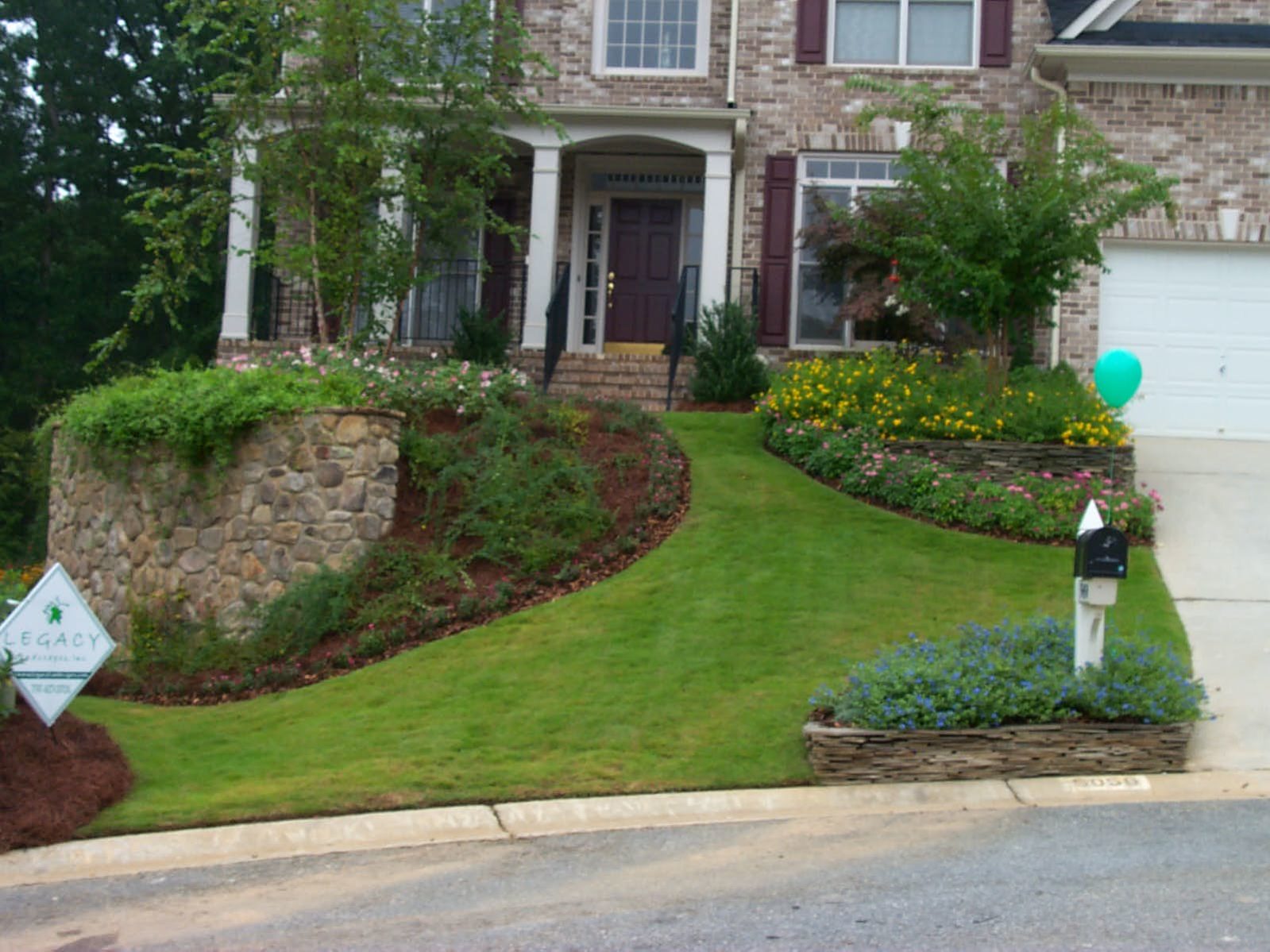 Brick house with landscaped front yard featuring flower beds, green lawn, a stone retaining wall, and a rustic deck overlooking it all. A blue balloon is tied to a mailbox, accompanied by a sign reading "Legacy" near the driveway.