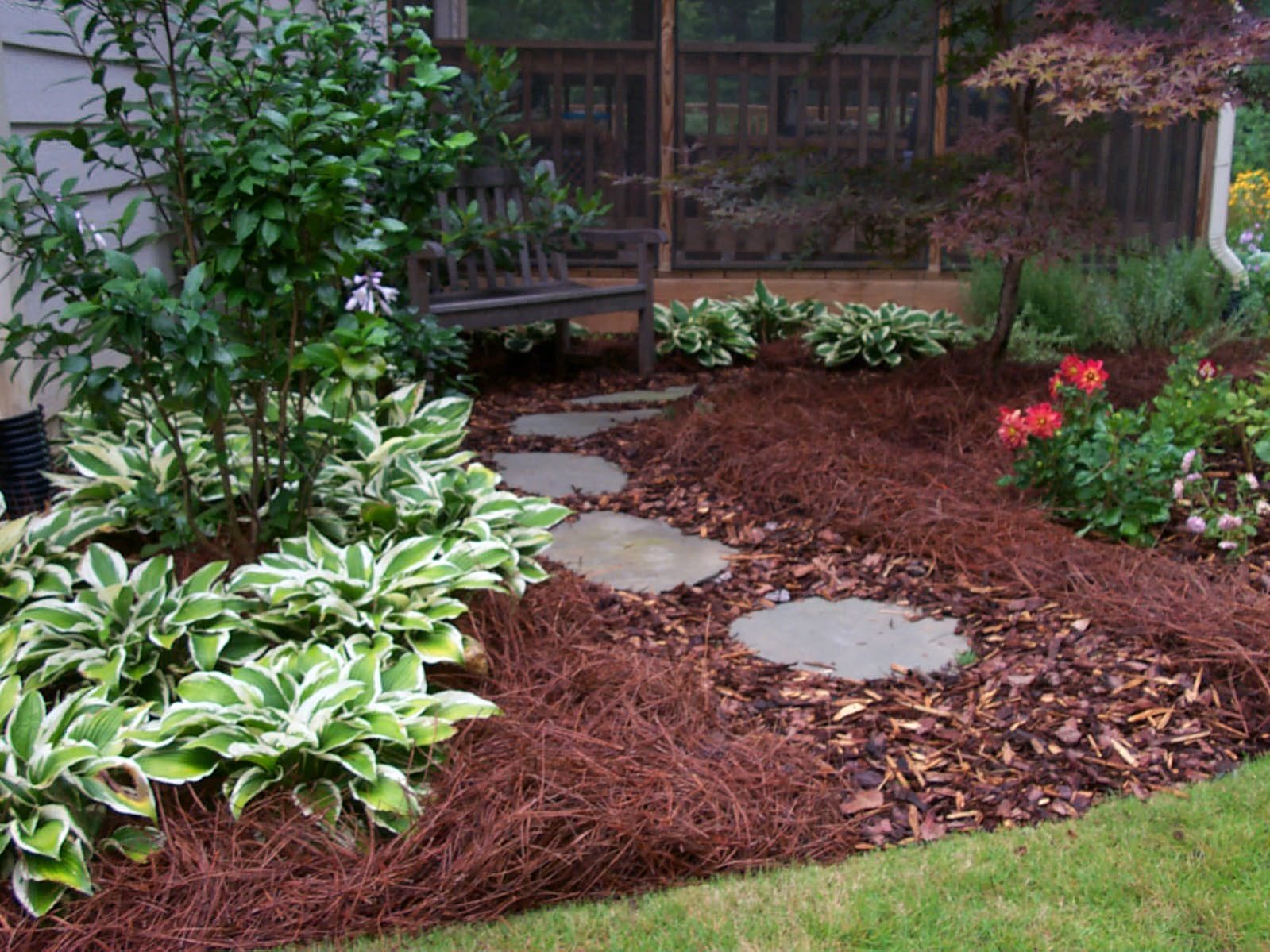 A garden with stepping stones, variegated hostas, and pine straw mulch complements the flowering plants near a screened porch and rustic deck.