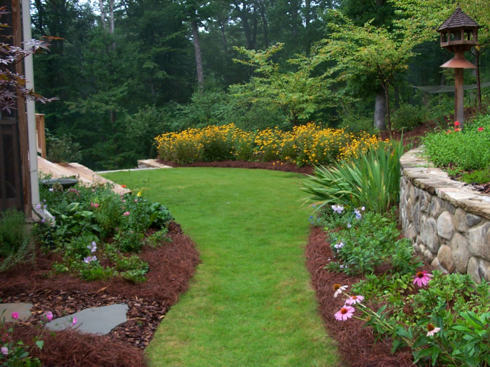 A well-maintained garden path lined with colorful flowers and shrubs leads to a rustic deck, bordered by a stone wall and trees in the background.