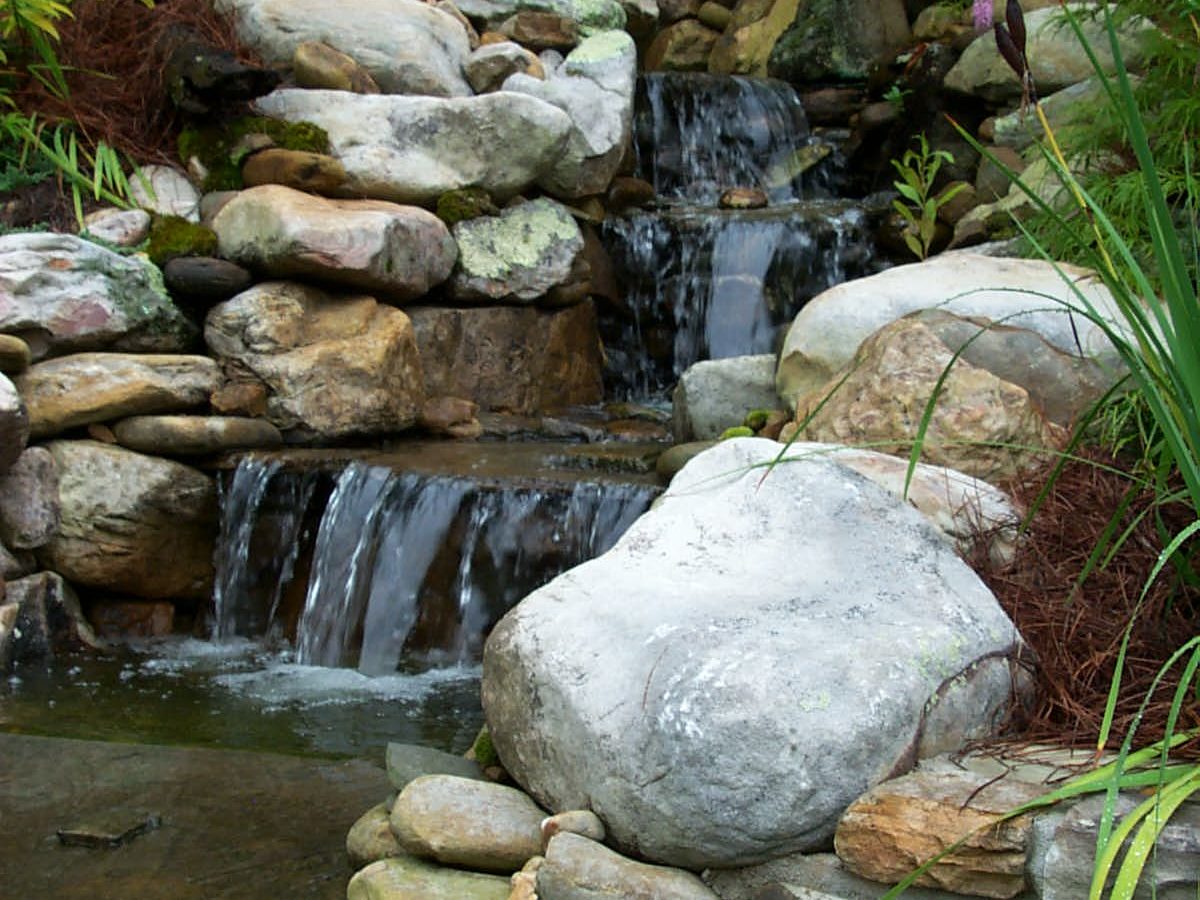 A rustic deck complements the artificial stone waterfall, surrounded by lush greenery.