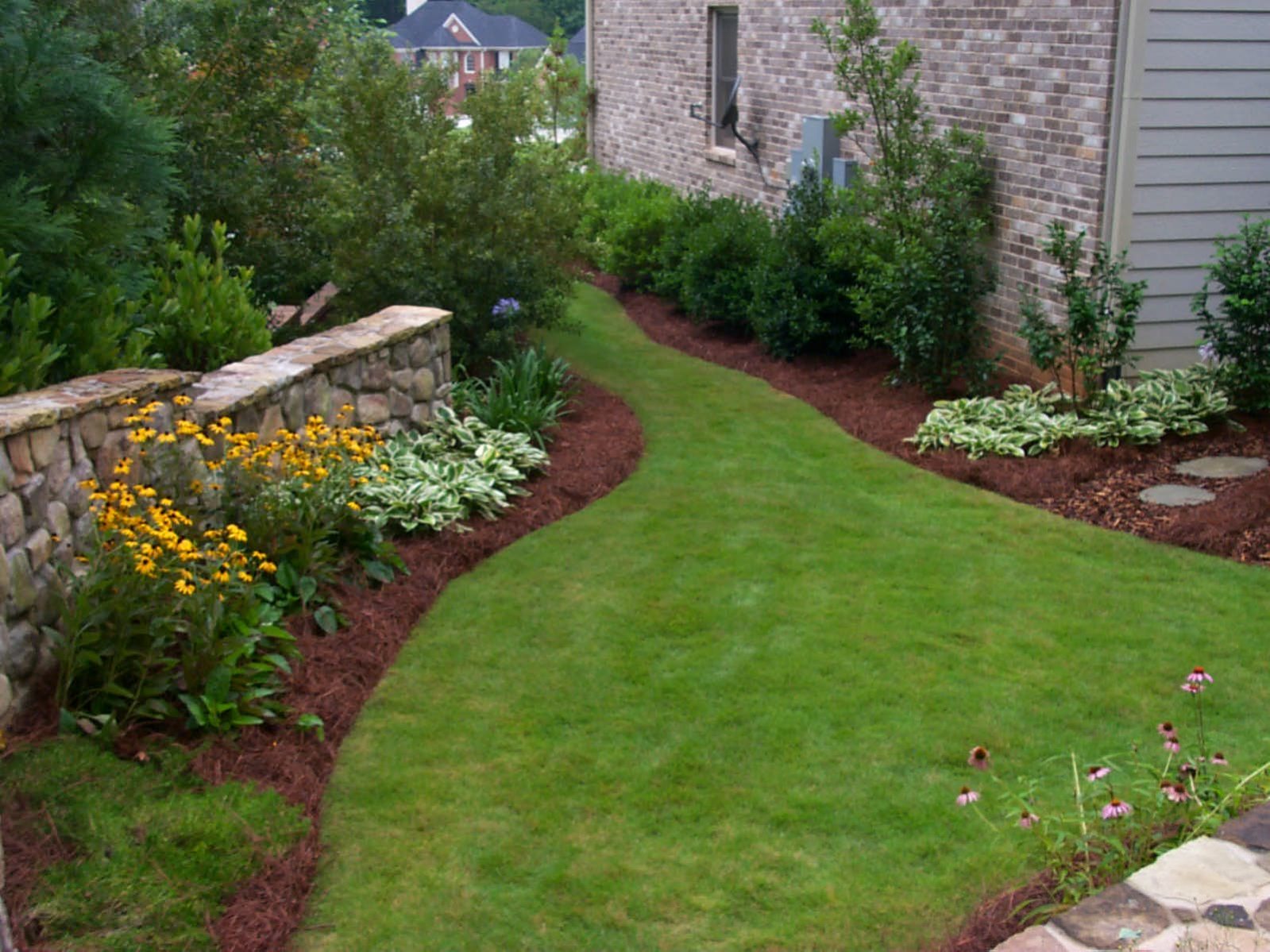 A narrow grass path curves between a stone wall and a house, flanked by flower beds and shrubs, leading to a rustic deck that invites relaxation.