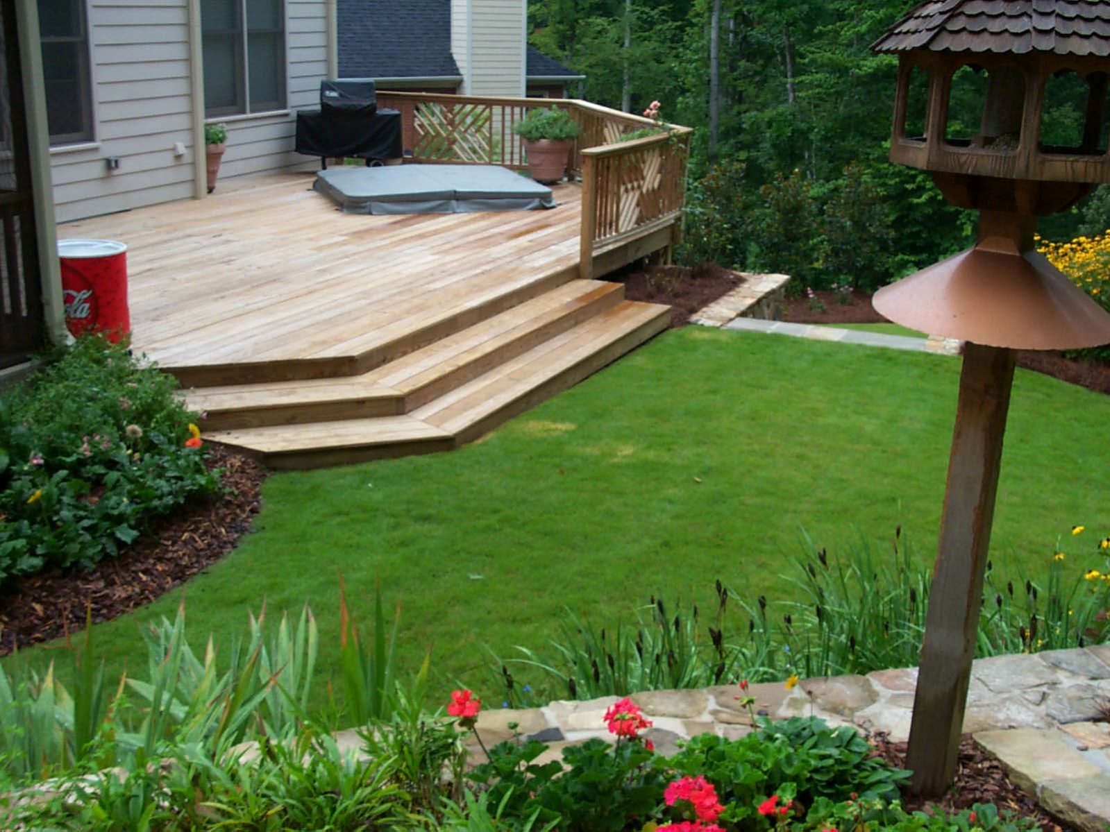 Rustic deck with steps, featuring a hot tub, potted plants, and a birdhouse overlooking a well-maintained grassy yard surrounded by vibrant flower beds.