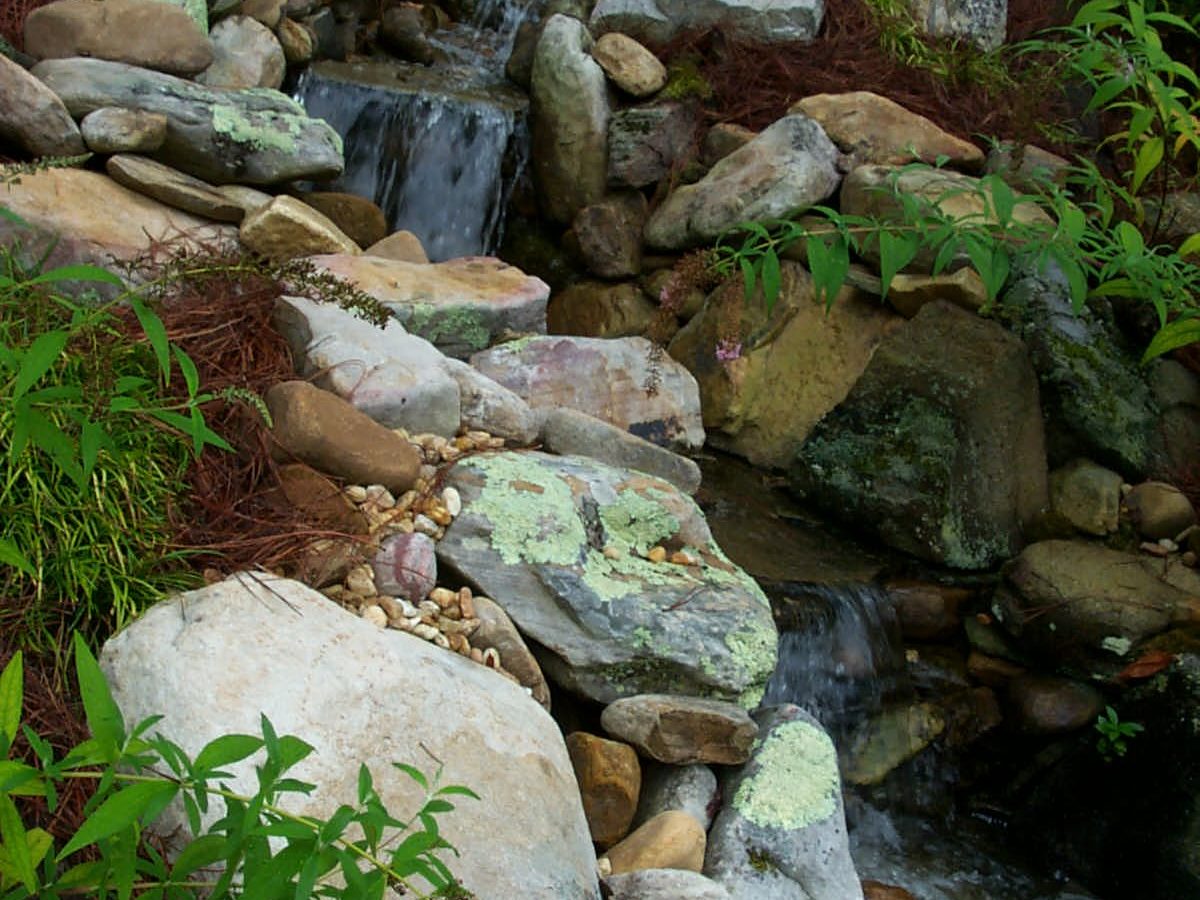 A small cascading waterfall in a garden flows over stacked rocks, surrounded by lush green plants. Nearby, a rustic deck provides the perfect spot to relax and enjoy the soothing sounds of nature.