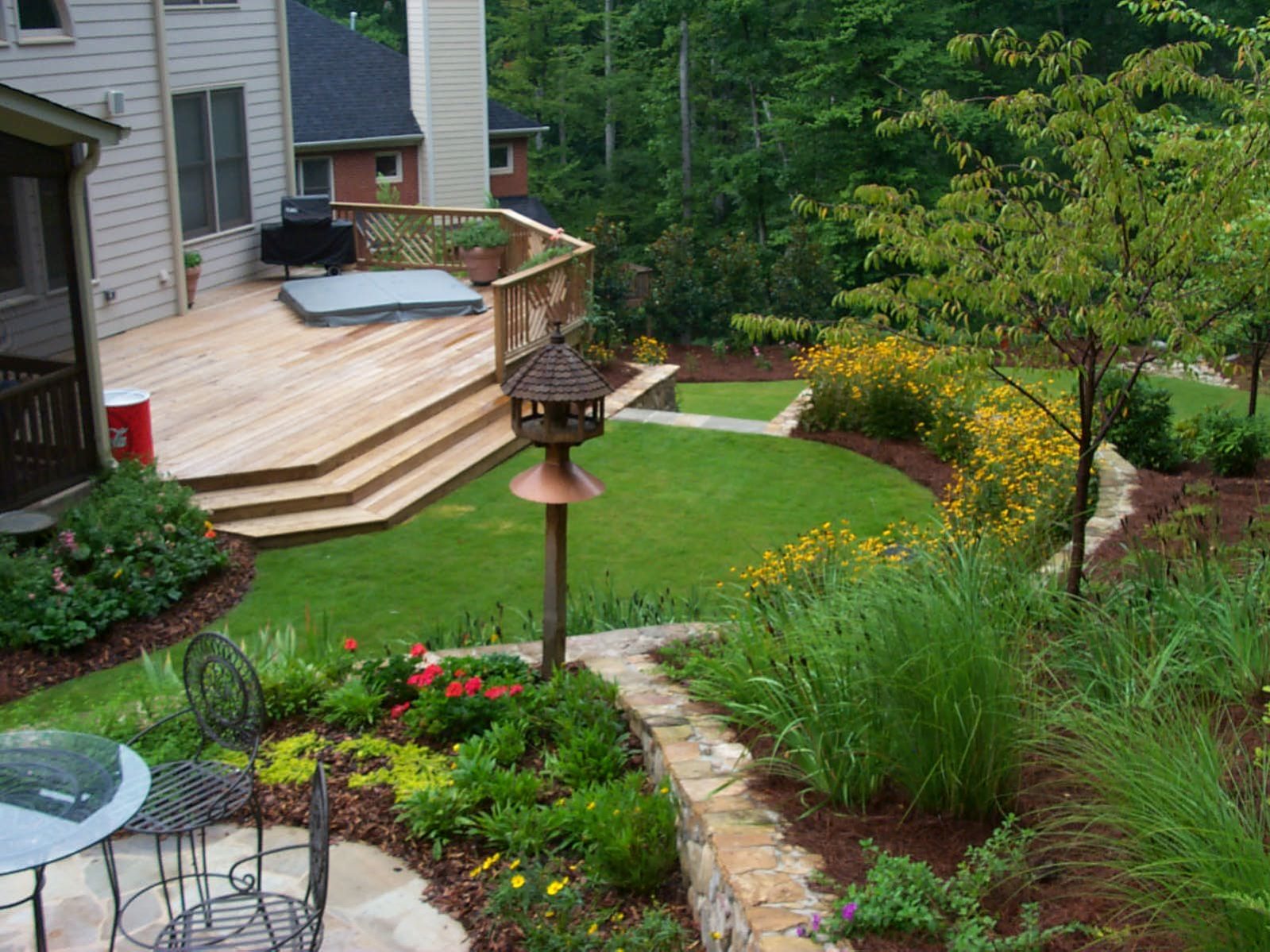 A landscaped backyard features a rustic deck overlooking a curved lawn, flowerbeds, and a stone path. Nearby, a metal patio table set nestles beside a lamppost, inviting you to relax in this charming outdoor space.