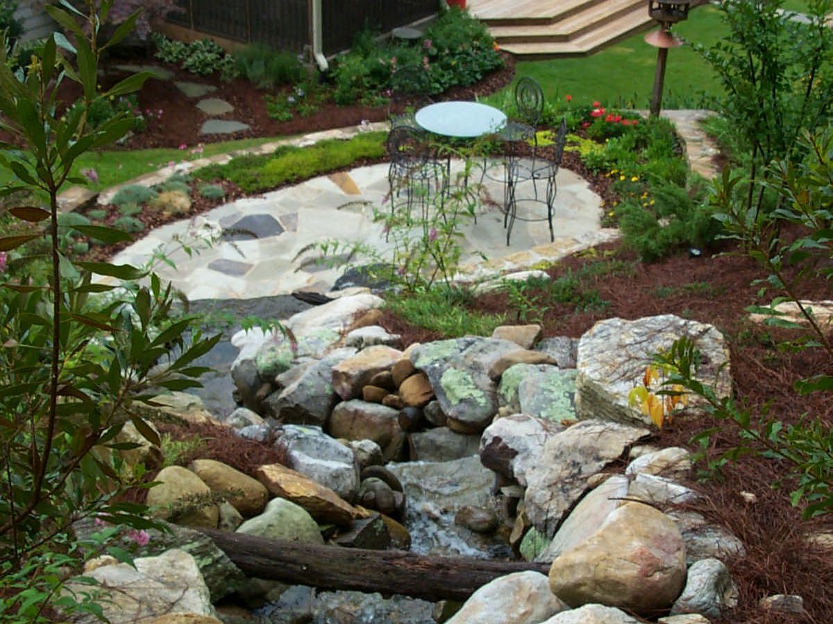 The backyard features a rustic deck with a hot tub, complemented by a small stream flowing over rocks. A patio with a table and chairs invites relaxation amid the lush greenery.
