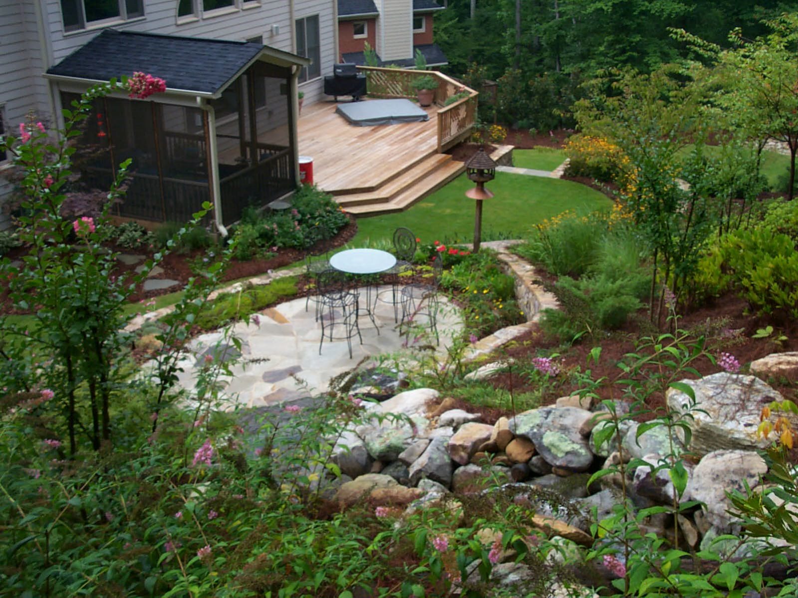 A backyard oasis with a rustic deck, complete with a patio set and stone path. The lush landscaping features rocks, grass, and vibrant flowering plants, creating an inviting retreat.