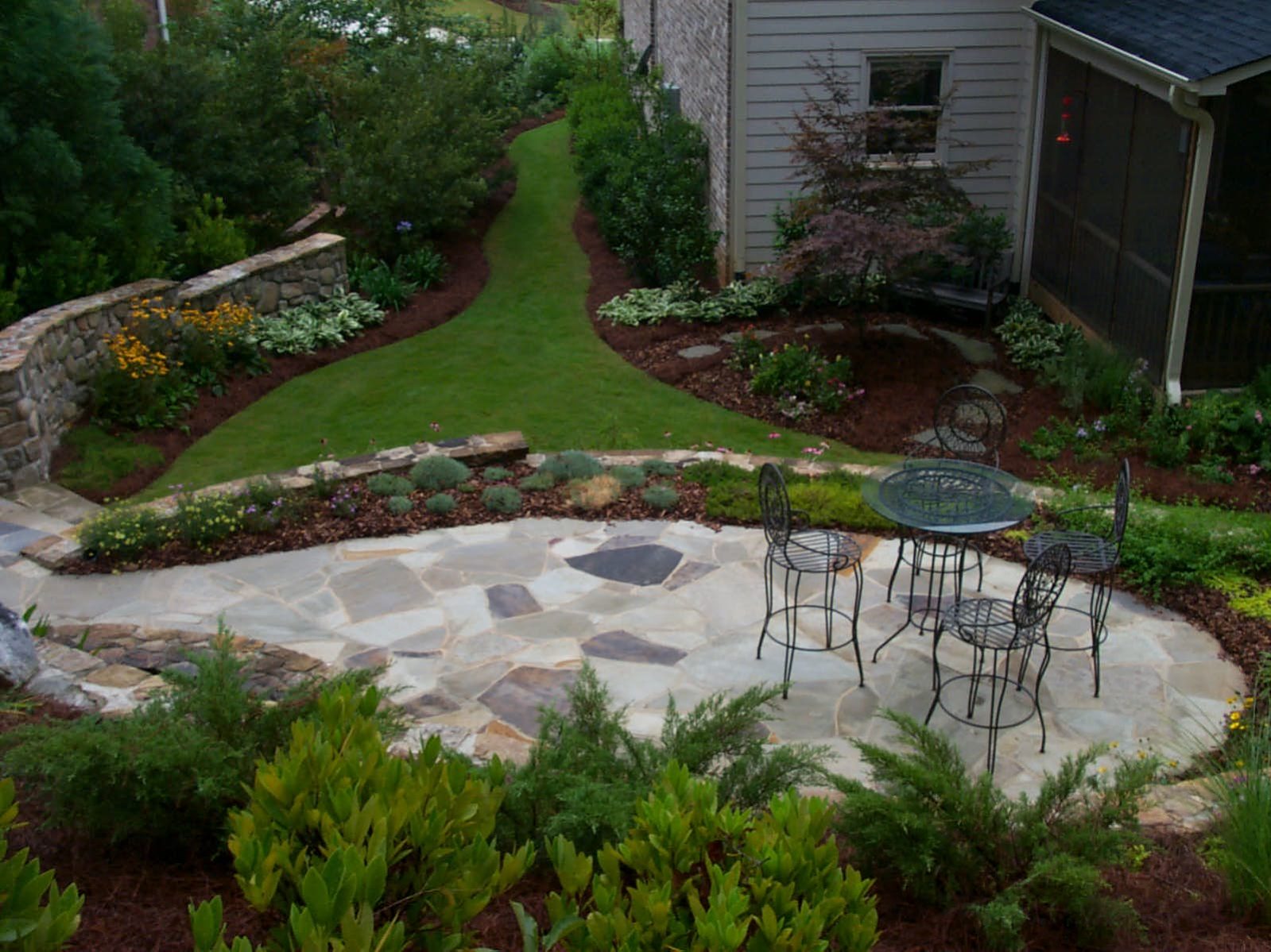 A rustic deck leads to a stone patio where a metal table and chairs await, surrounded by a garden with pathways, lush greenery, and timeless stone walls.