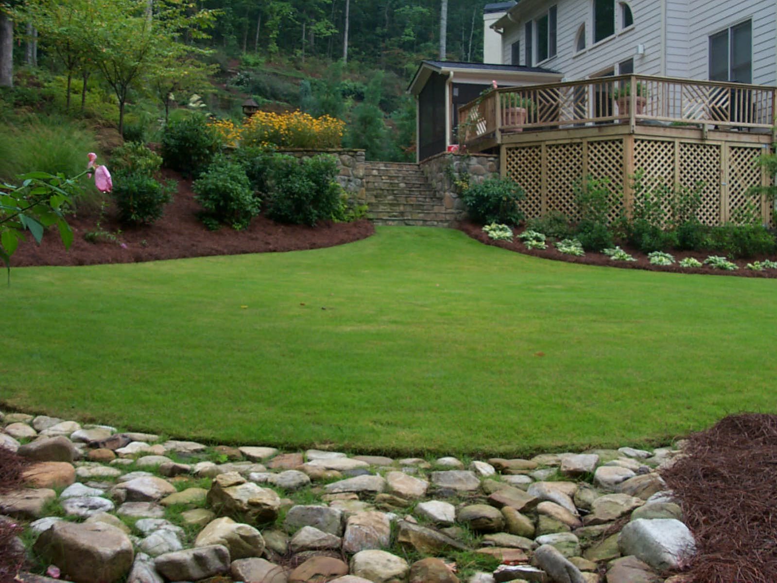 A landscaped backyard features a green lawn, bordered by shrubs and flowers. A rustic wooden deck extends from a house on the right, and stones form a dry creek bed in the foreground.