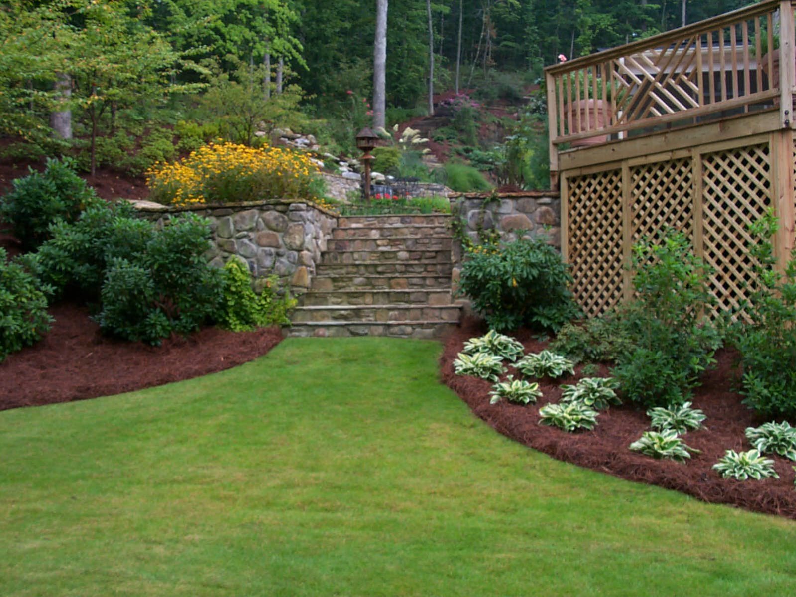 A well-maintained garden with a green lawn, stone steps, and vibrant flower beds showcases a rustic deck adorned with lattice detail. In the background, trees and shrubs offer an inviting canopy of greenery.
