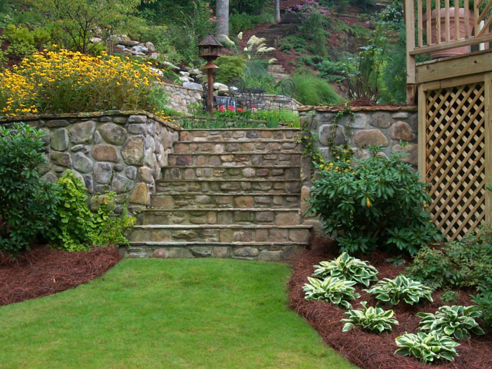 A garden with stone steps, surrounded by lush greenery, shrubs, and flowers, leads to a raised area featuring a rustic deck with a wooden structure.
