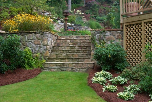 A garden with stone steps, surrounded by lush greenery, shrubs, and flowers, leads to a raised area featuring a rustic deck with a wooden structure.