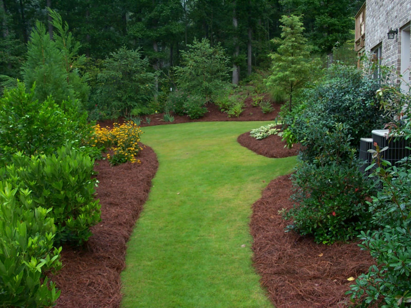 A well-maintained backyard with a green grass path, bordered by lush trees and shrubbery, leads to a rustic deck nestled in the wooded area.