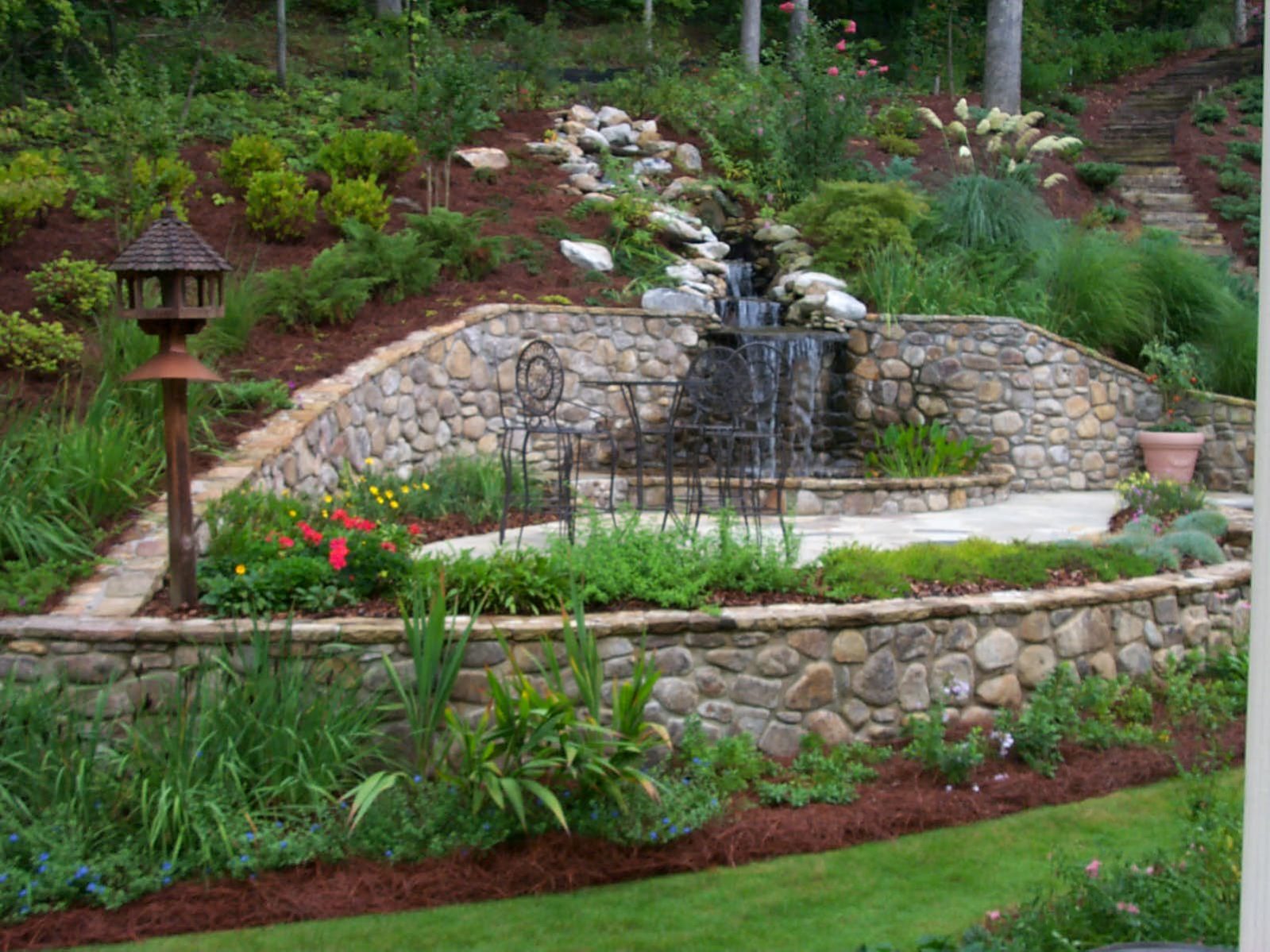 A landscaped garden with a stone retaining wall features a small waterfall and metal chairs set against the backdrop of lush greenery, complemented by a rustic deck that adds charm to the serene oasis.
