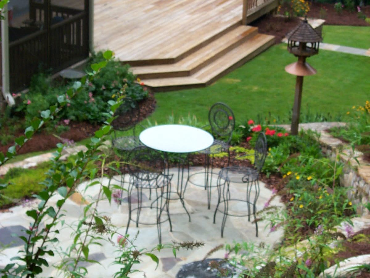 A backyard featuring a rustic deck, stone patio, wire chairs, and a small table, complemented by a hot tub surrounded by lush plants and greenery.