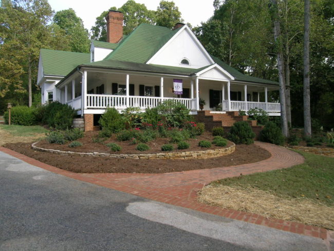 This charming country farmhouse boasts a large white exterior and a green roof, complemented by a wraparound porch. It's beautifully surrounded by landscaped garden beds and mature trees, with a welcoming brick pathway leading to the entrance.