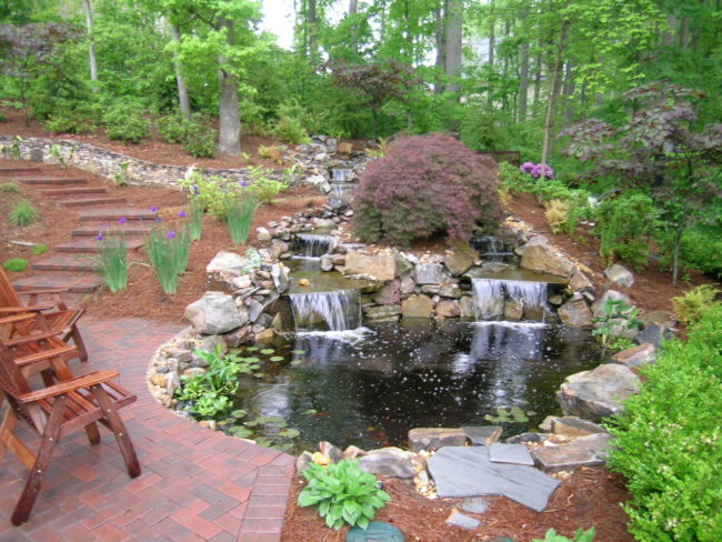 A serene garden scene features a tranquil pond with small waterfalls, surrounded by lush greenery. On the left, a brick path guides you to wooden chairs, perfect for relaxing by the water’s edge. A serene garden scene features a tranquil pond with small waterfalls, surrounded by lush greenery. On the left, a brick path guides you to wooden chairs, perfect for relaxing by the water's edge.
