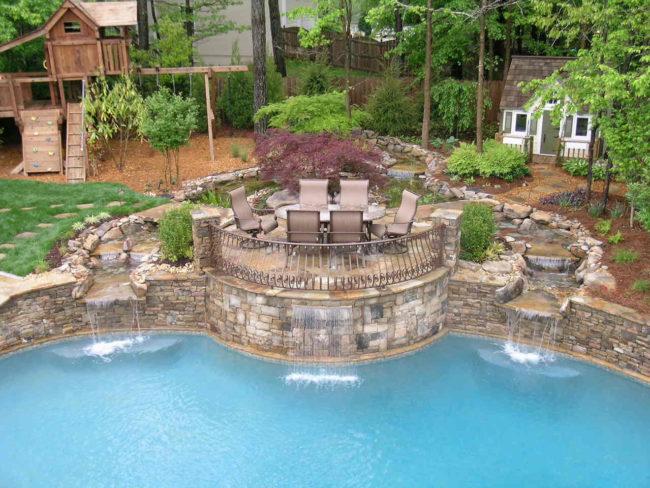 A rustic pool graces the backyard scene, complemented by a stone waterfall feature. Nearby, a patio set rests on a raised area, overlooking a wooden play structure and a quaint shed nestled among trees and shrubs.