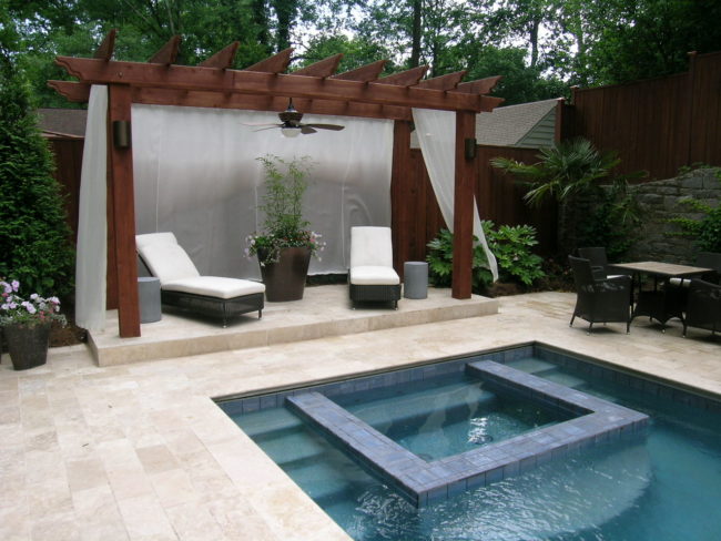 The outdoor poolside area features a wooden pergola with white curtains, two lounge chairs, and a potted plant. Nearby, find a table with chairs for relaxing by the pool and spa. The pool boasts a square inset section surrounded by elegant stone tiles.