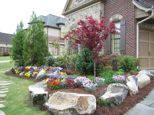 A well-maintained garden with colorful flowers, small shrubs, and a red-leafed tree seamlessly integrated into the landscaping. Surrounded by boulders, it sits elegantly in front of a charming brick house.