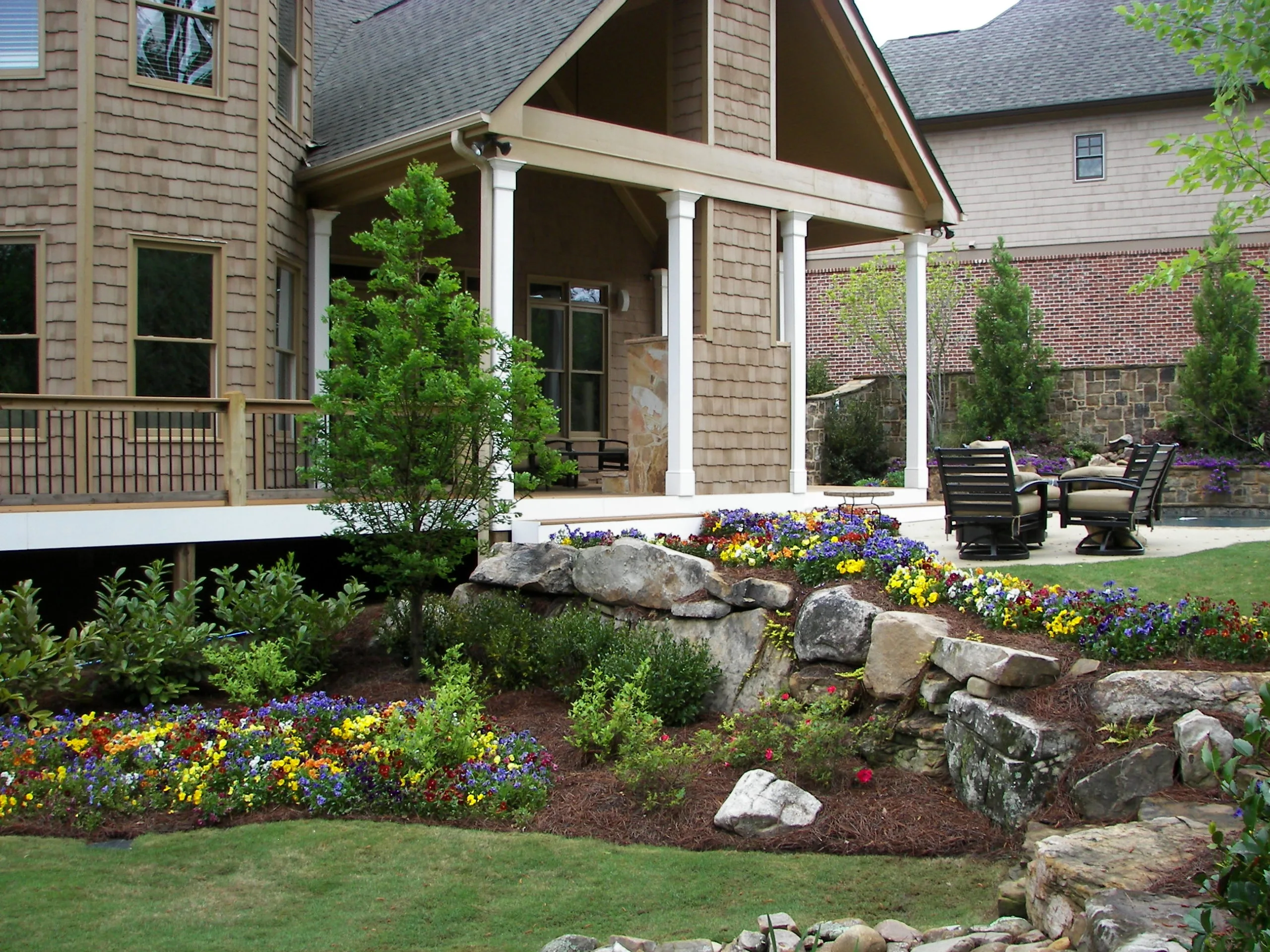 A well-maintained backyard garden with a vibrant landscape of colorful flowers, stone features, and a patio furnished for relaxation, nestled near a house with a charming covered porch. A well-maintained backyard garden with a vibrant landscape of colorful flowers, stone features, and a patio furnished for relaxation, nestled near a house with a charming covered porch.