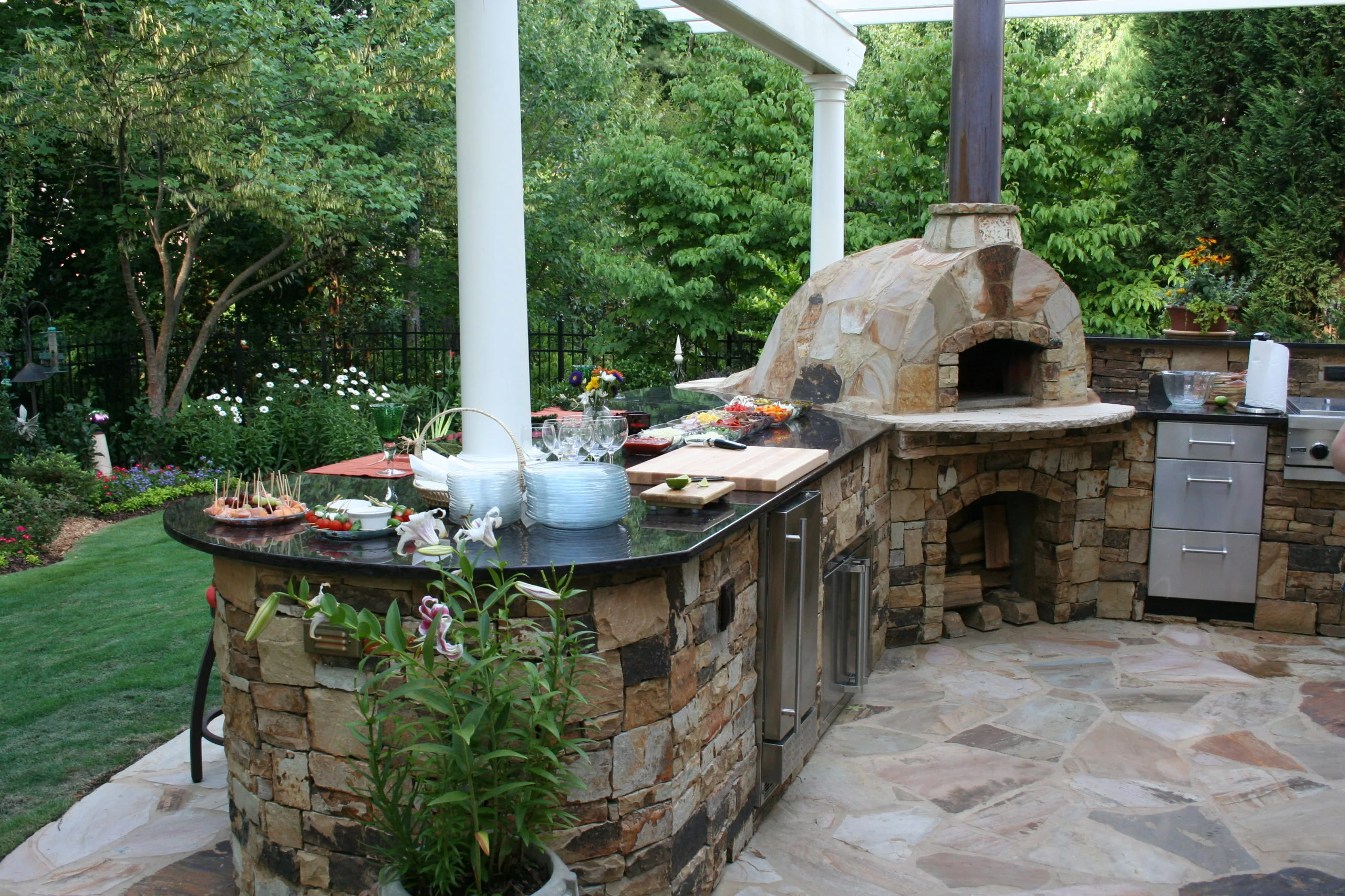 An outside kitchen complete with a stone pizza oven, a countertop laden with food and drinks, all embraced by lush greenery. An outside kitchen complete with a stone pizza oven, a countertop laden with food and drinks, all embraced by lush greenery.