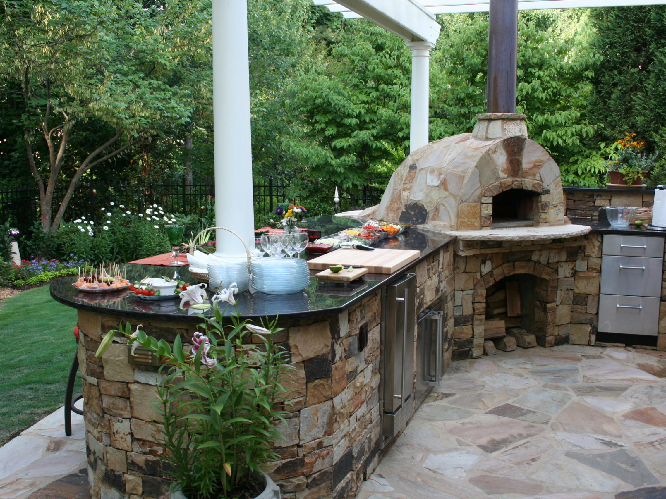 An outdoor kitchen featuring a stone pizza oven and a cozy fireplace, with a countertop full of dishes and appetizers, surrounded by lush greenery.