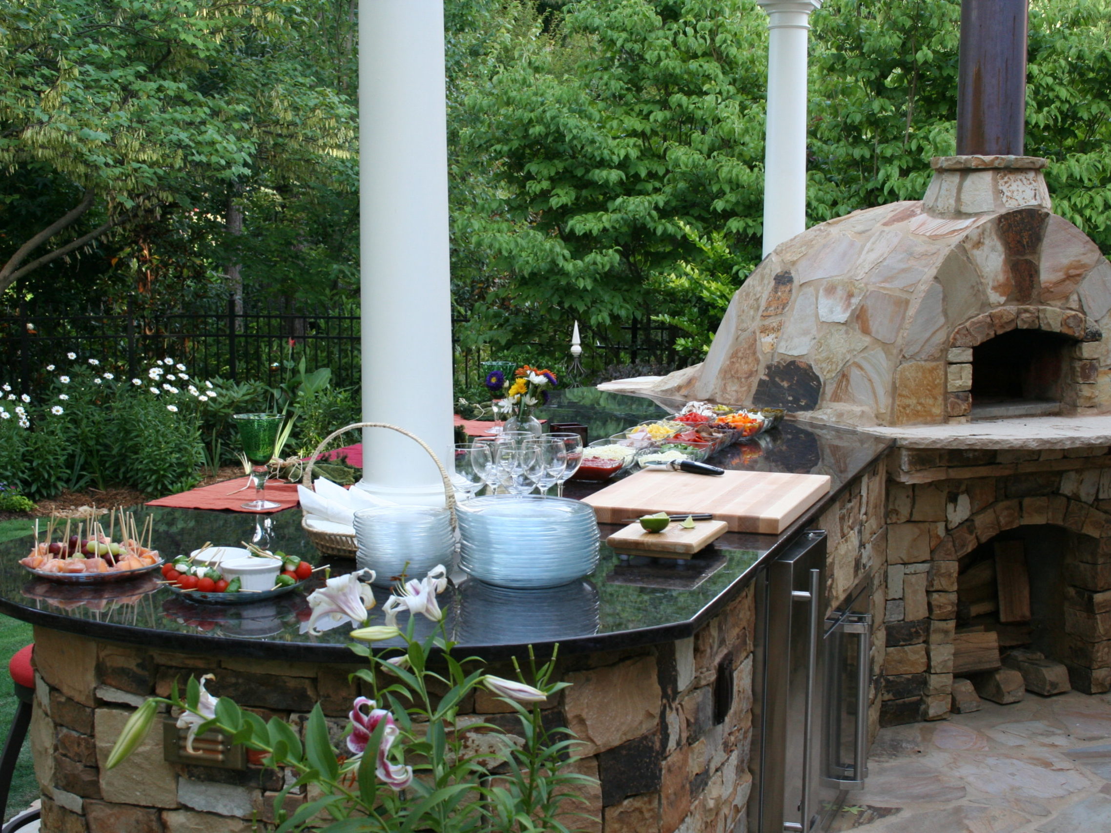 An outdoor kitchen features a stone pizza oven and a cozy fireplace, alongside a countertop adorned with plates, utensils, and neatly arranged appetizers. The setting is beautifully surrounded by lush greenery and vibrant flowers.