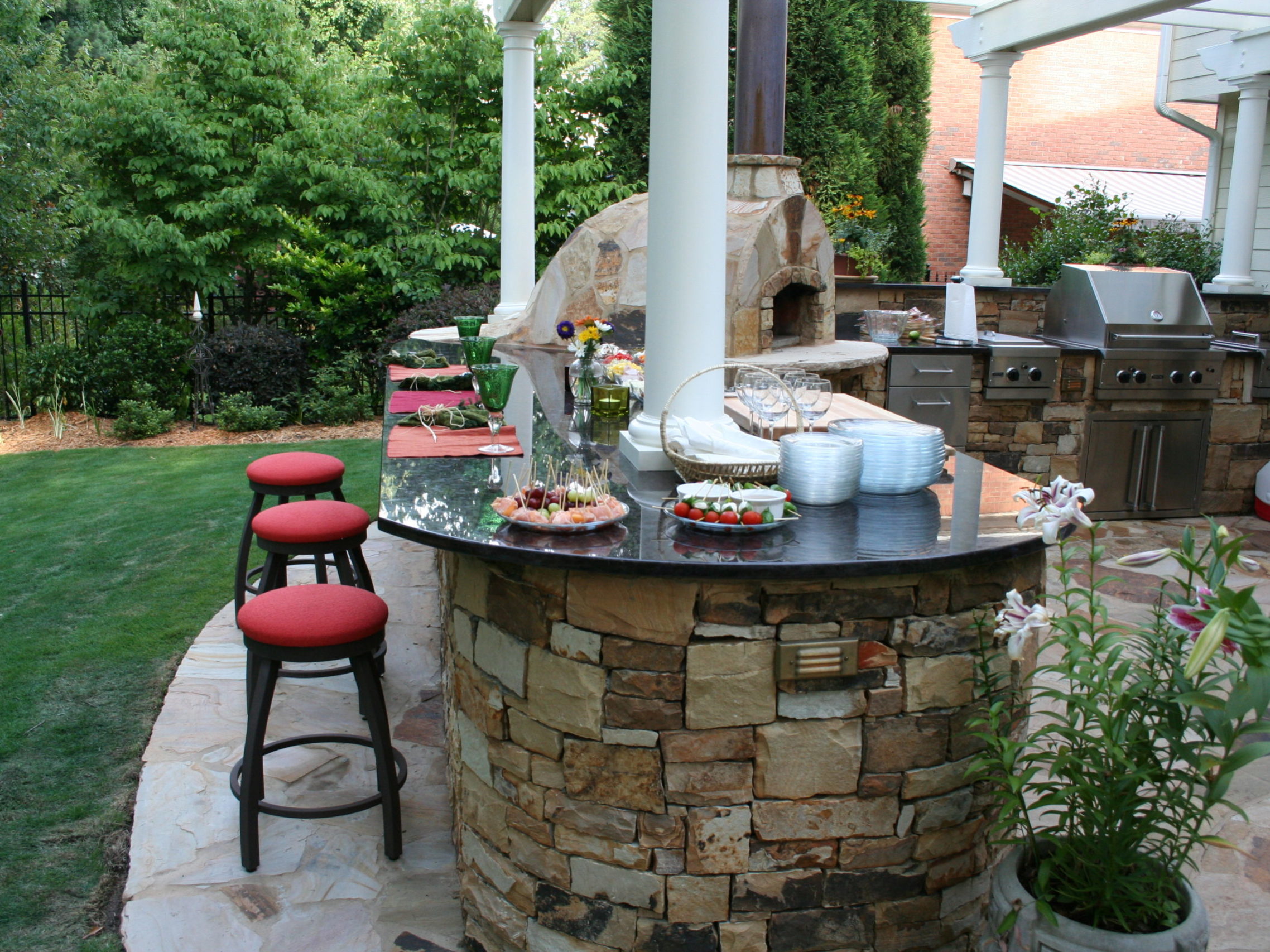 Outdoor kitchen with a stone bar and grill area, featuring a cozy fireplace and barstools with red cushions. Plates of food sit beside the pizza oven while lush greenery surrounds the patio.