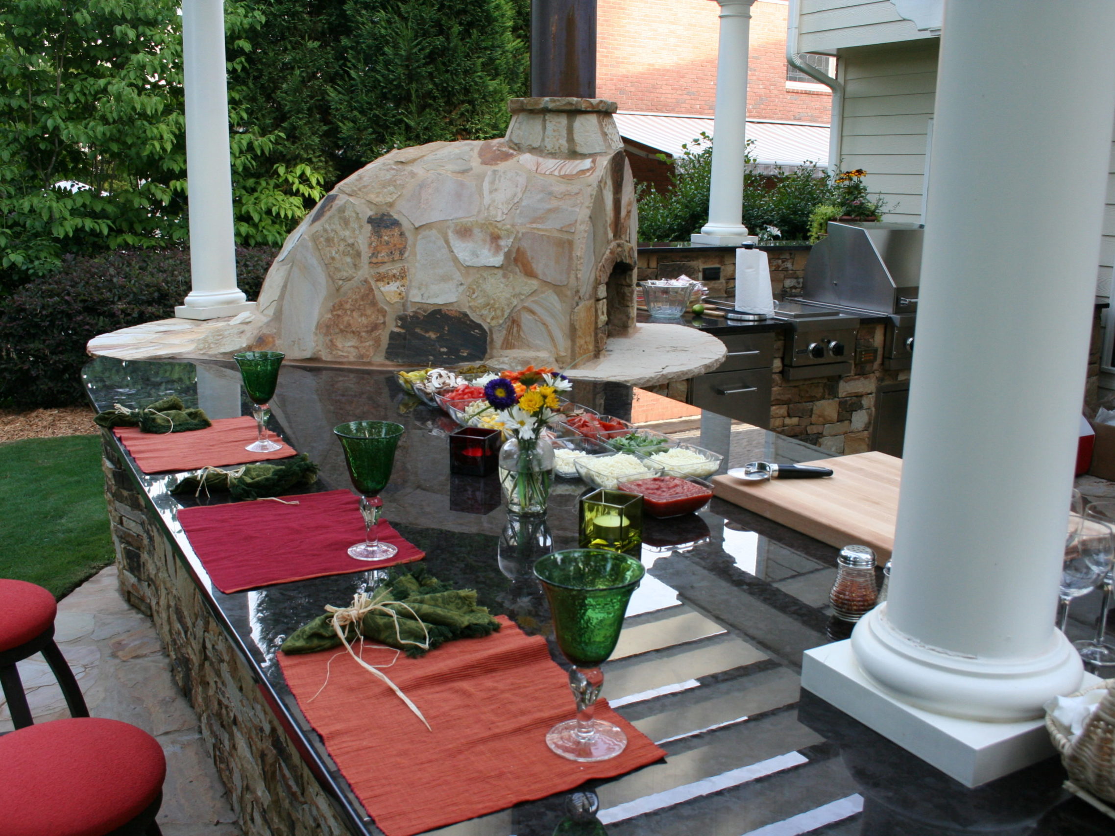 Outdoor kitchen featuring a stone pizza oven and fireplace, a granite counter set with red and orange placemats, green glasses, and vibrant floral centerpieces. White columns frame the scene with a grill subtly tucked in the background.