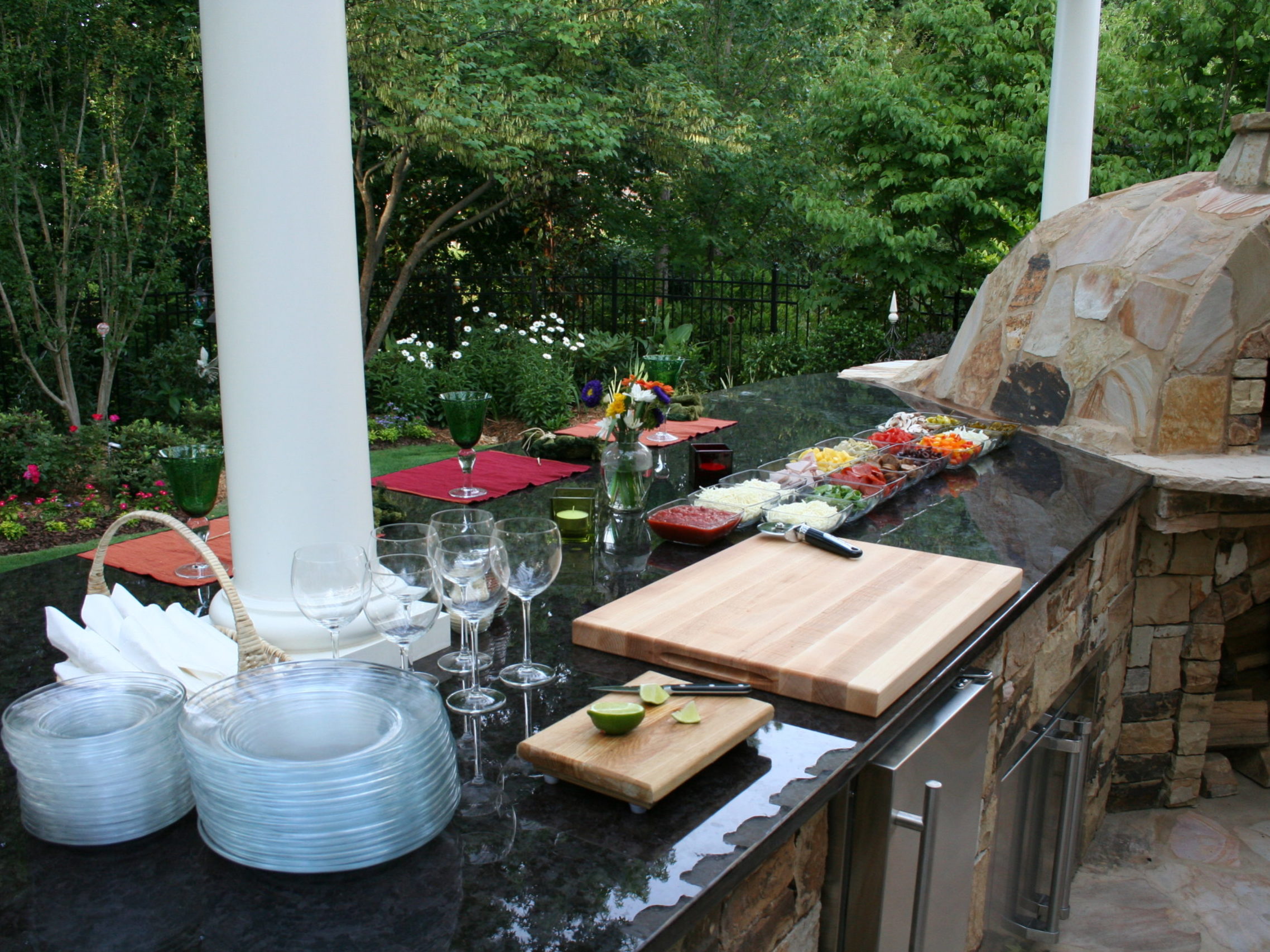 An outdoor kitchen with a stone pizza oven and fireplace features cutting boards, glassware, and a variety of prepared ingredients on a granite countertop. Lush greenery surrounds the area, offering a perfect blend of functionality and natural beauty.