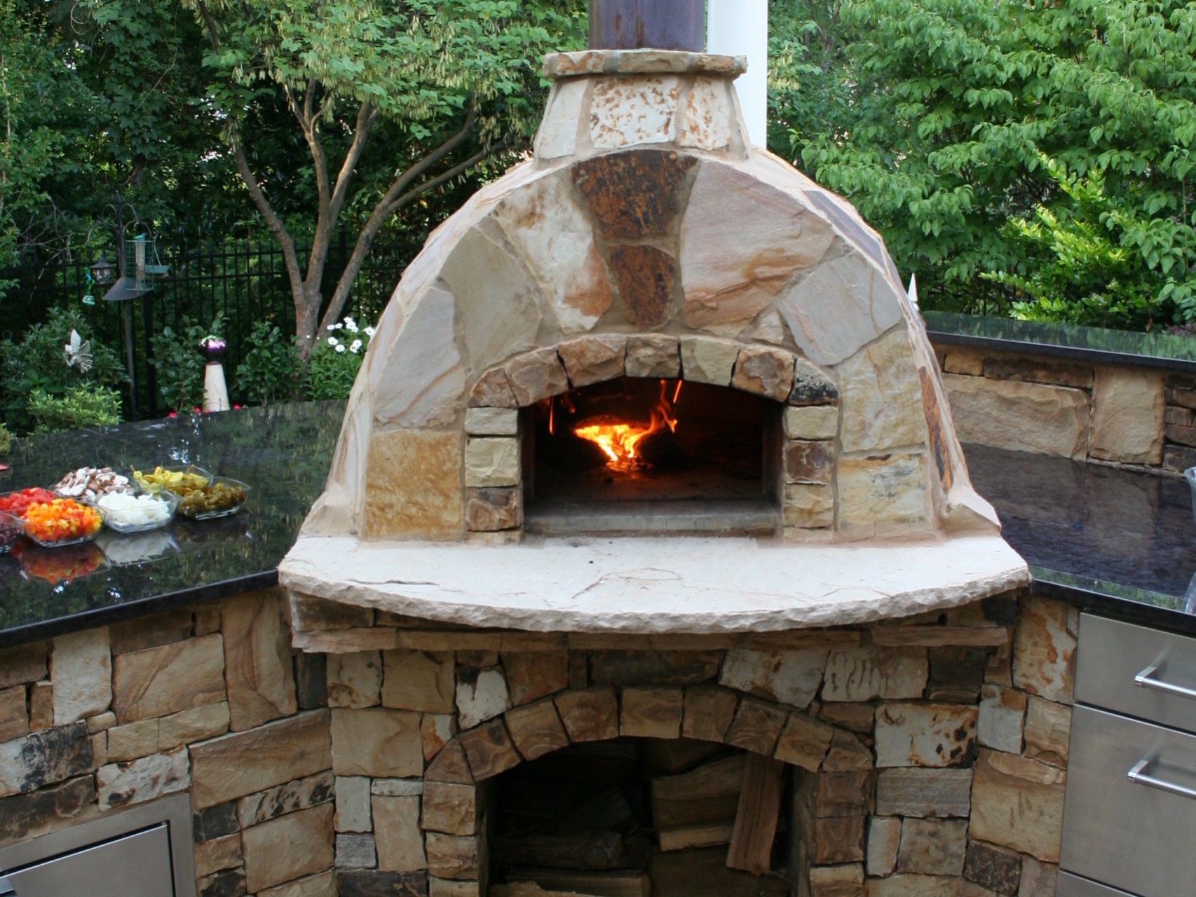 Outdoor stone pizza oven with fire burning, seamlessly integrated into an elegant outdoor kitchen and fireplace setup. Surrounded by countertops and cabinets, all nestled in a charming garden setting with logs neatly stored underneath.