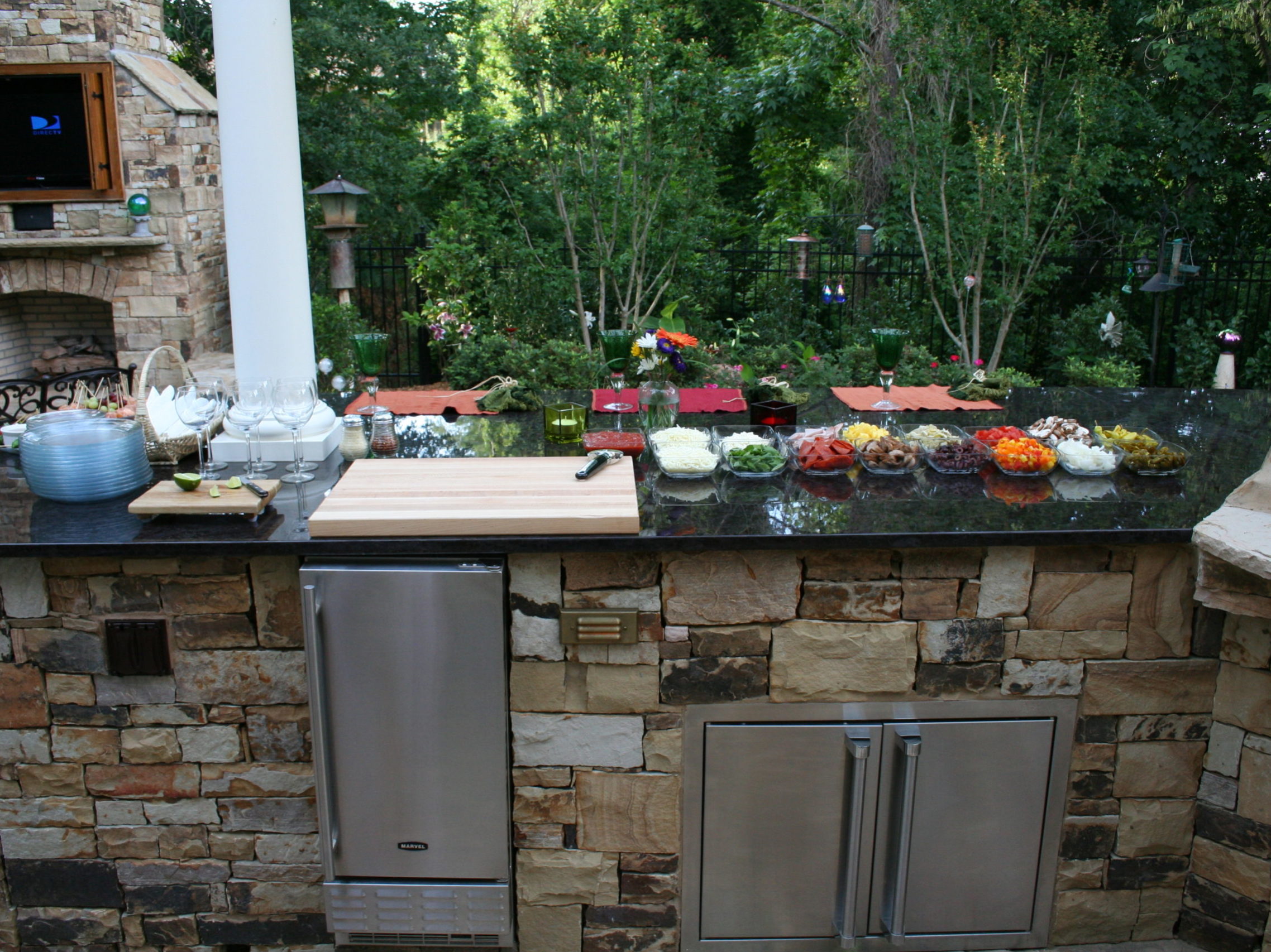 Outdoor kitchen featuring stone countertops, various dishes and ingredients, a built-in grill, and a sink. A cozy outdoor fireplace complements the setting, all surrounded by trees and lush greenery.