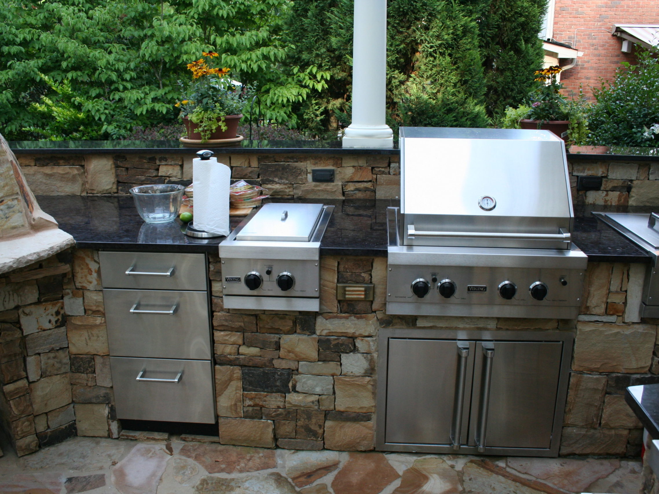 An outdoor stone kitchen featuring stainless steel appliances, including a grill and cabinets, is complemented by a cozy fireplace, all set against a serene backdrop of trees.