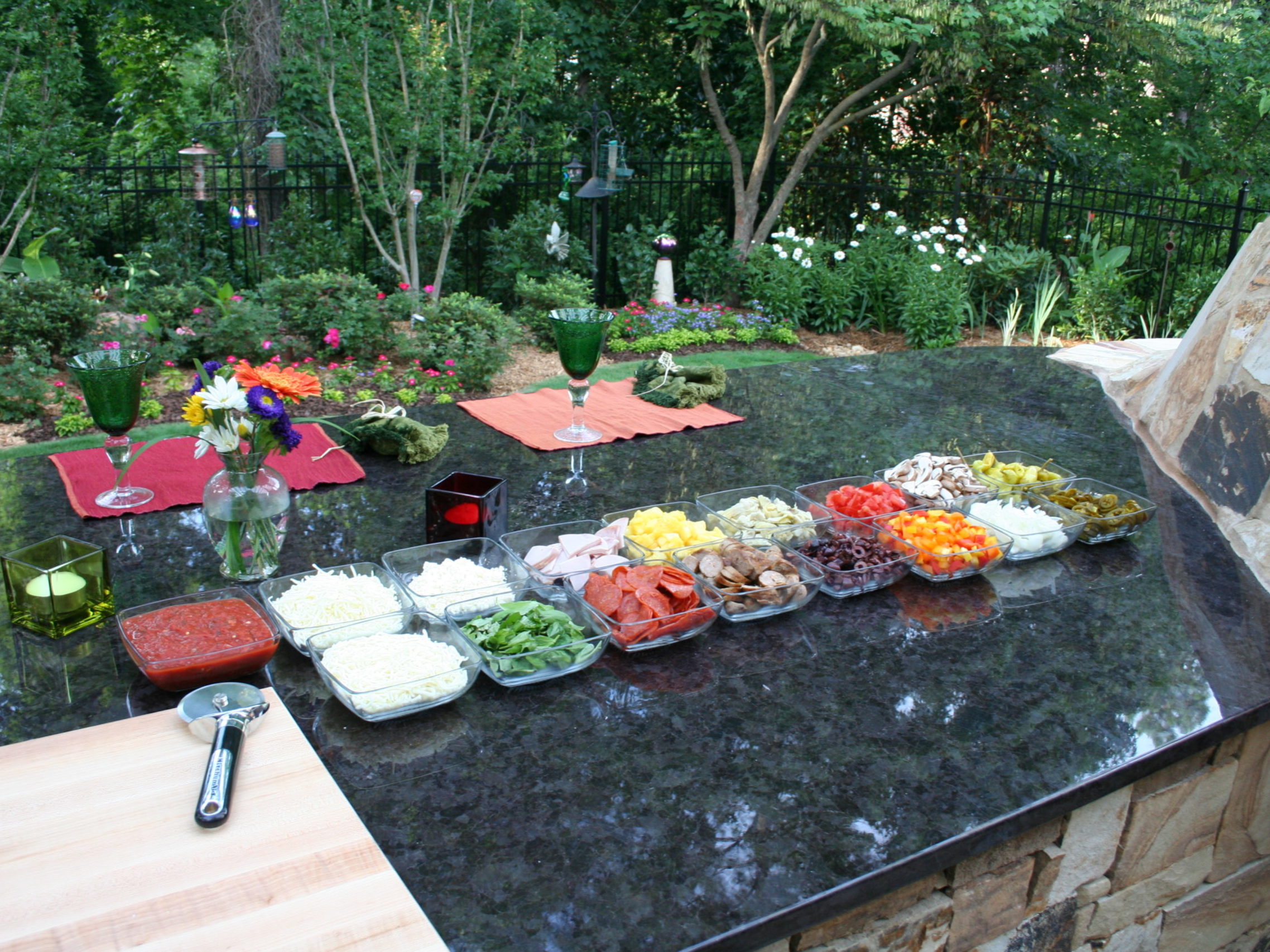 Outdoor kitchen setup with a variety of toppings in small glass dishes on a stone countertop. A cozy fireplace warms the scene, while the lush garden in the background blooms with vibrant flowers and trees.