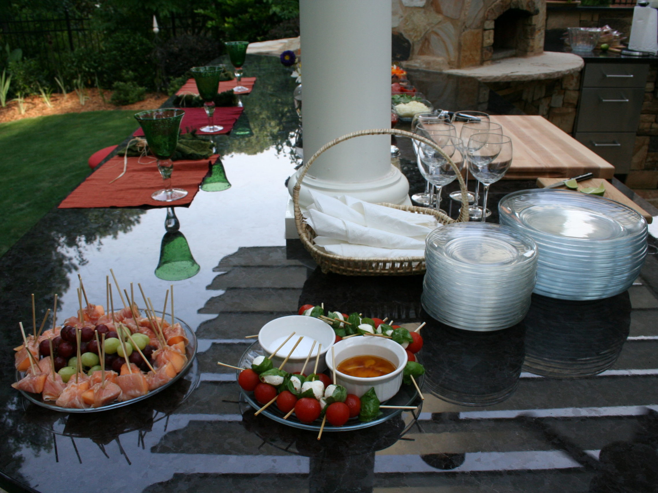 Outdoor table setting with skewered appetizers, a basket with napkins, glasses, and plates. A grill and countertop are in the background, complemented by an inviting outdoor kitchen and fireplace that promise warmth and delightful gatherings.