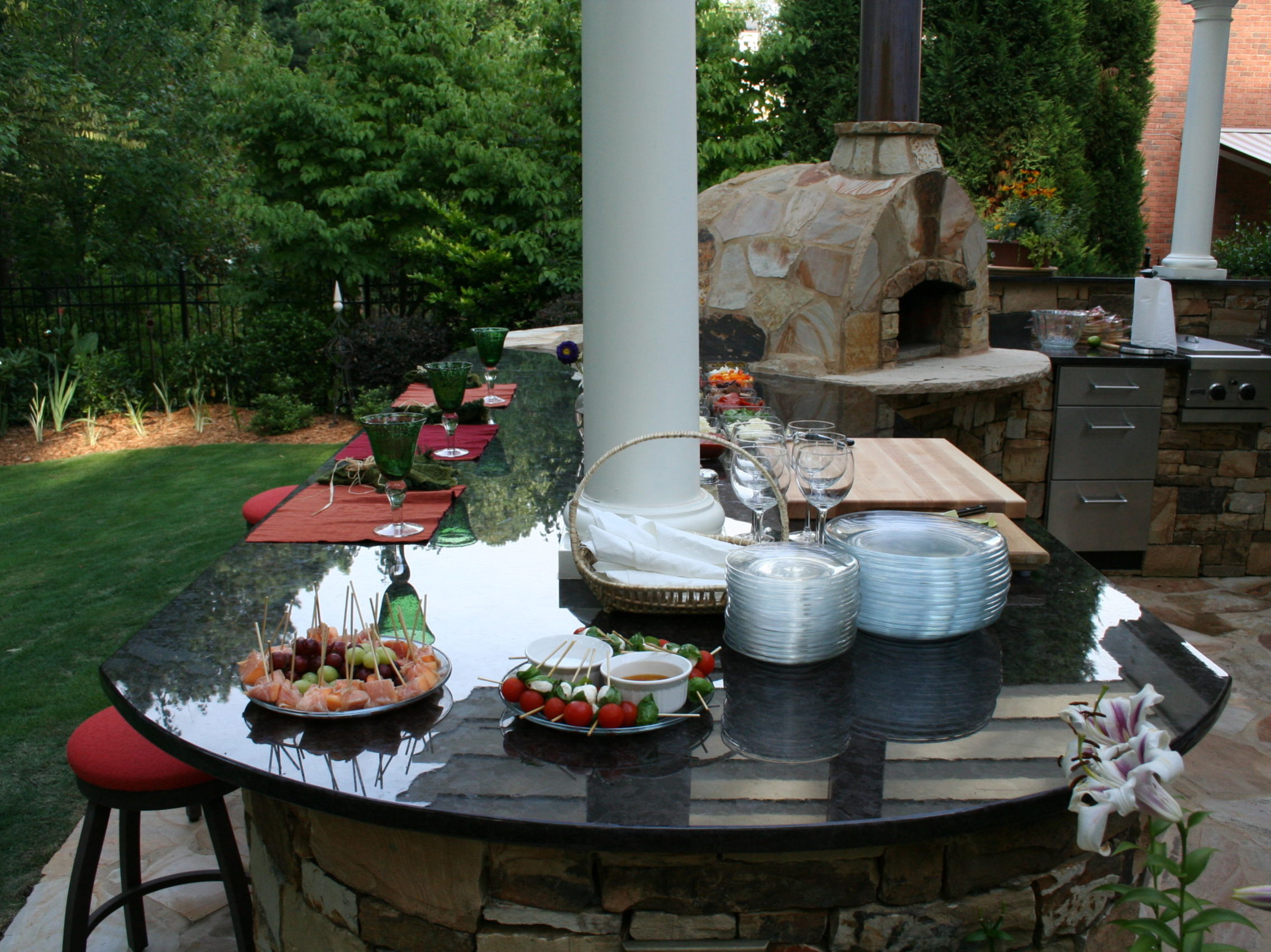 An outdoor kitchen featuring a stone pizza oven and a cozy fireplace, complemented by a granite countertop adorned with plates, glasses, and a platter of appetizers. This serene setting is surrounded by lush trees and grass.