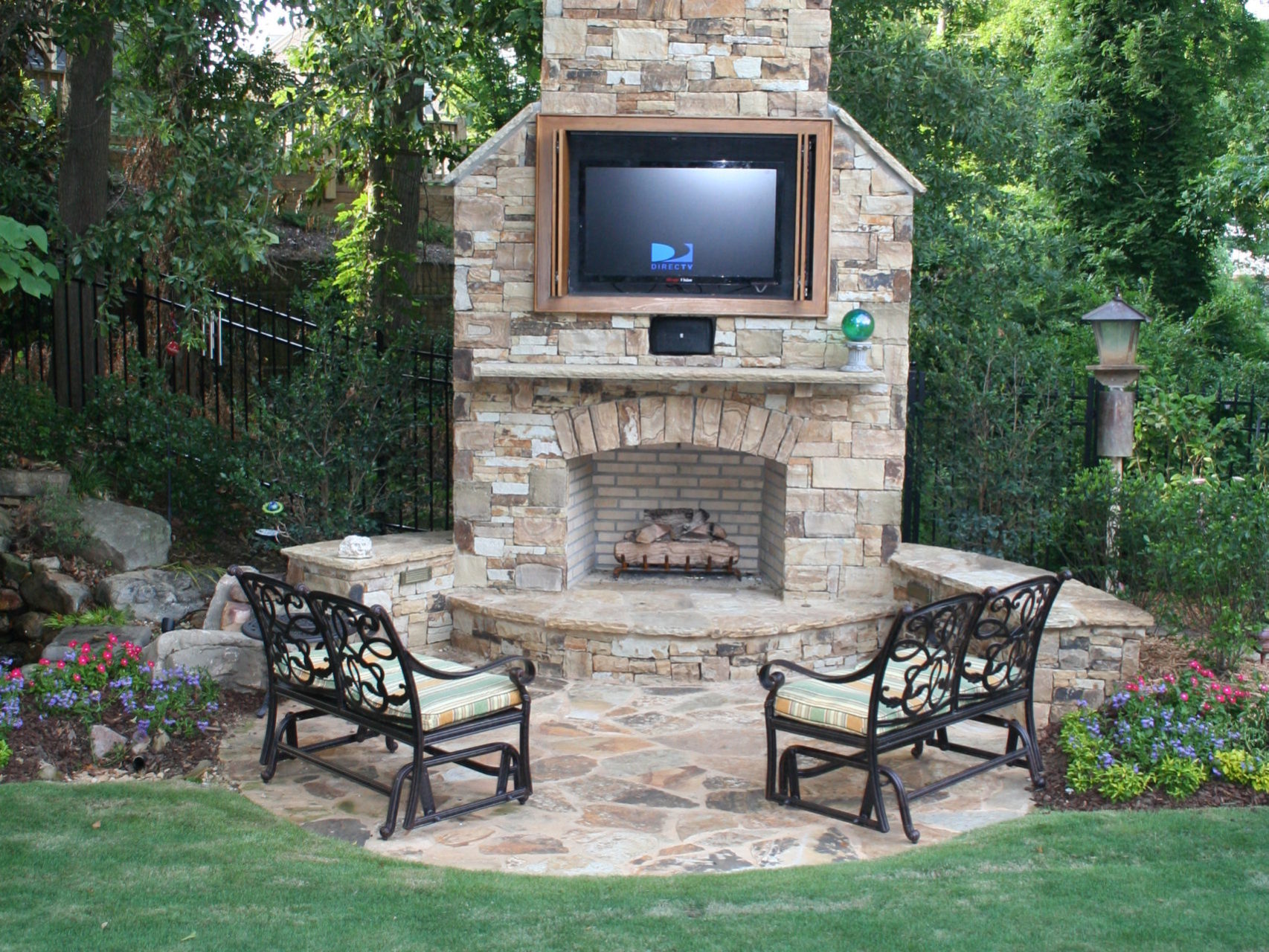 Outdoor stone fireplace with a mounted TV is nestled among trees, accompanied by two metal chairs and vibrant flowerbeds. This cozy nook seamlessly extends from the outdoor kitchen, creating the perfect blend of modern comfort and nature's charm.