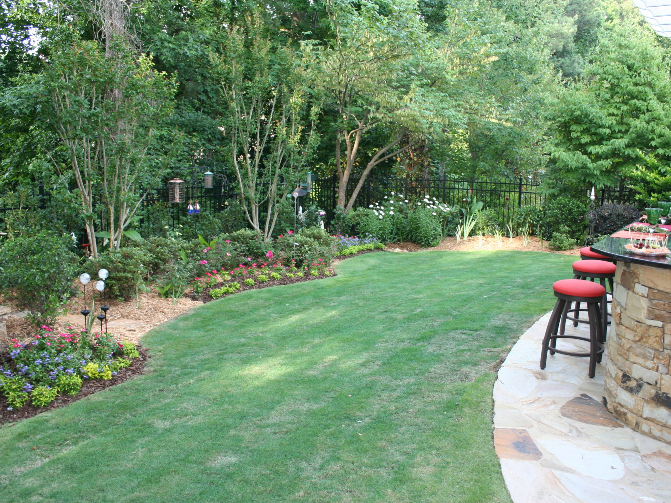 A well-maintained backyard featuring a stone bar with red stools, green grass, and flowering shrubs is complemented by an outdoor kitchen and fireplace nestled among tall trees.