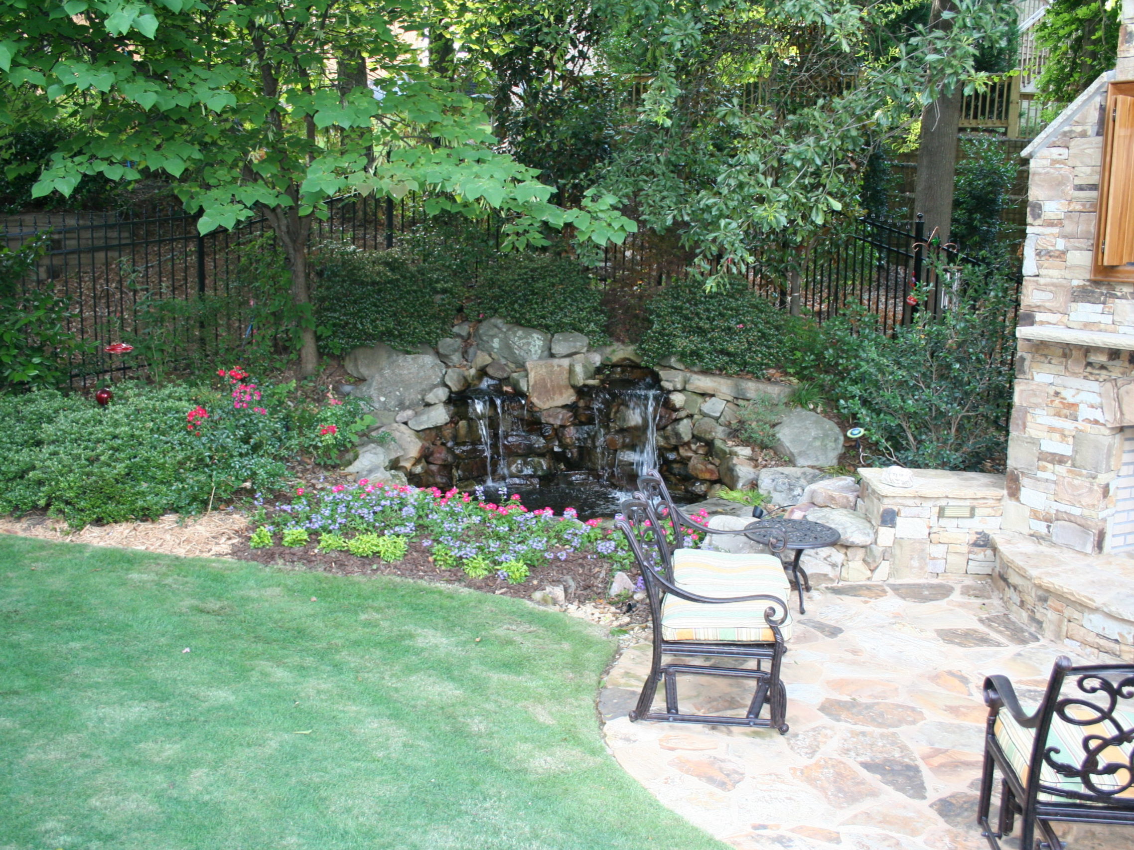 A landscaped backyard featuring a stone patio, outdoor kitchen, fireplace, small waterfall, garden bed brimming with flowers, and lush greenery.