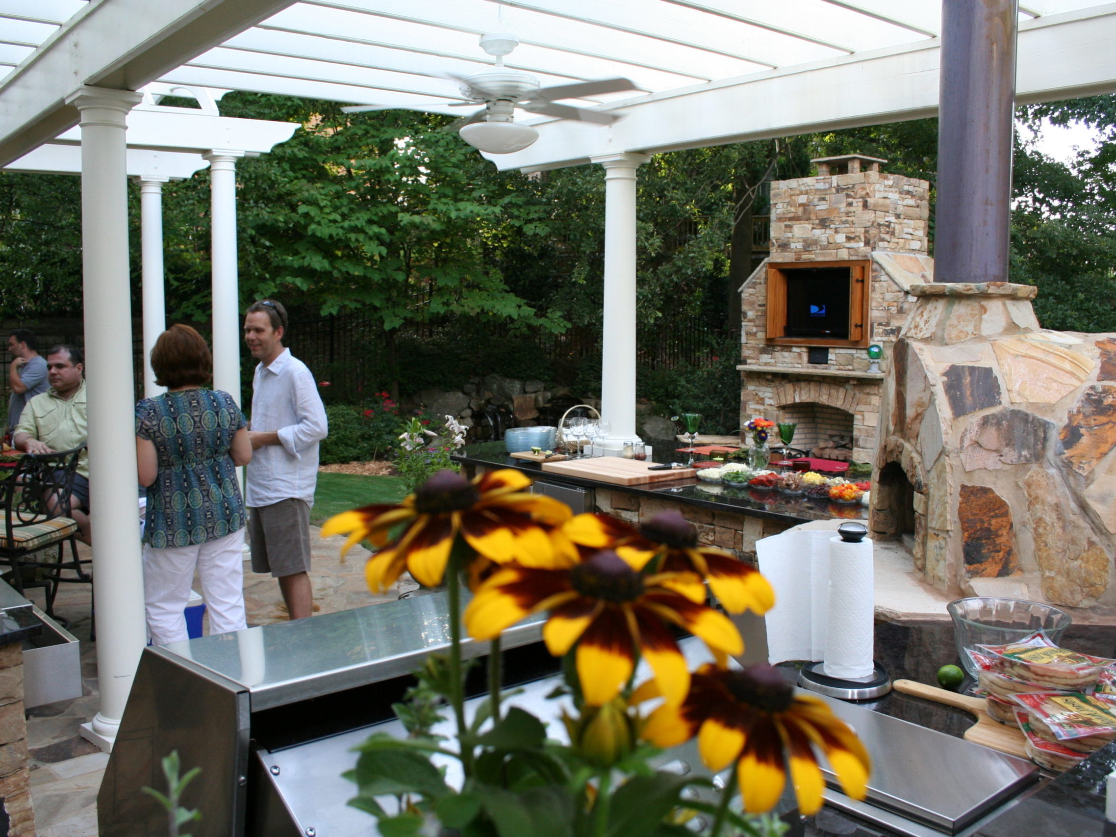 The outdoor kitchen boasts a stone pizza oven and a cozy fireplace, where people are happily chatting around a table laden with food. Bright yellow flowers add a cheerful touch to the scene.