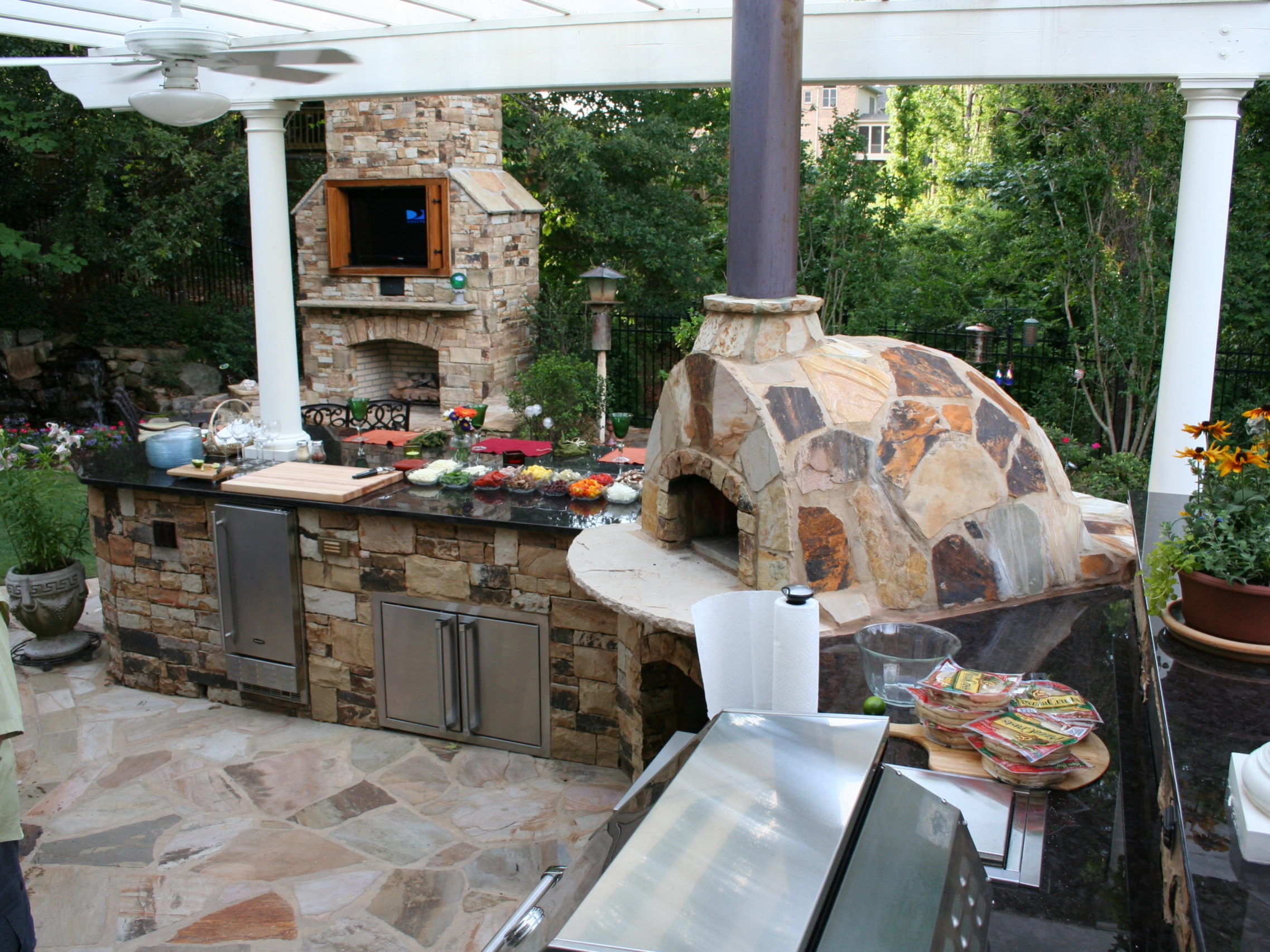 An outdoor kitchen boasts a stone pizza oven, grill, and countertops adorned with various dishes. Nestled in lush greenery, it offers a cozy ambiance with a partial view of the fireplace.