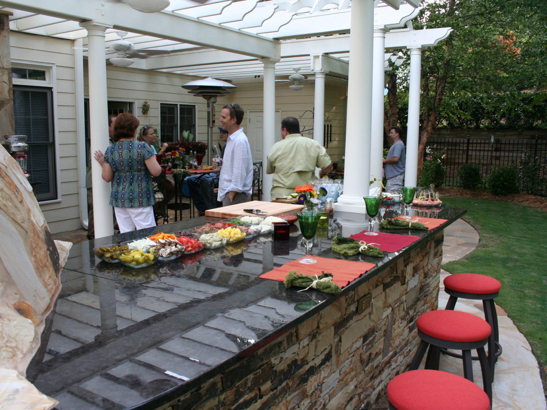 Outdoor gathering with people chatting near a stone bar counter with food and drinks, under a pergola in an inviting backyard featuring an outdoor kitchen and fireplace.