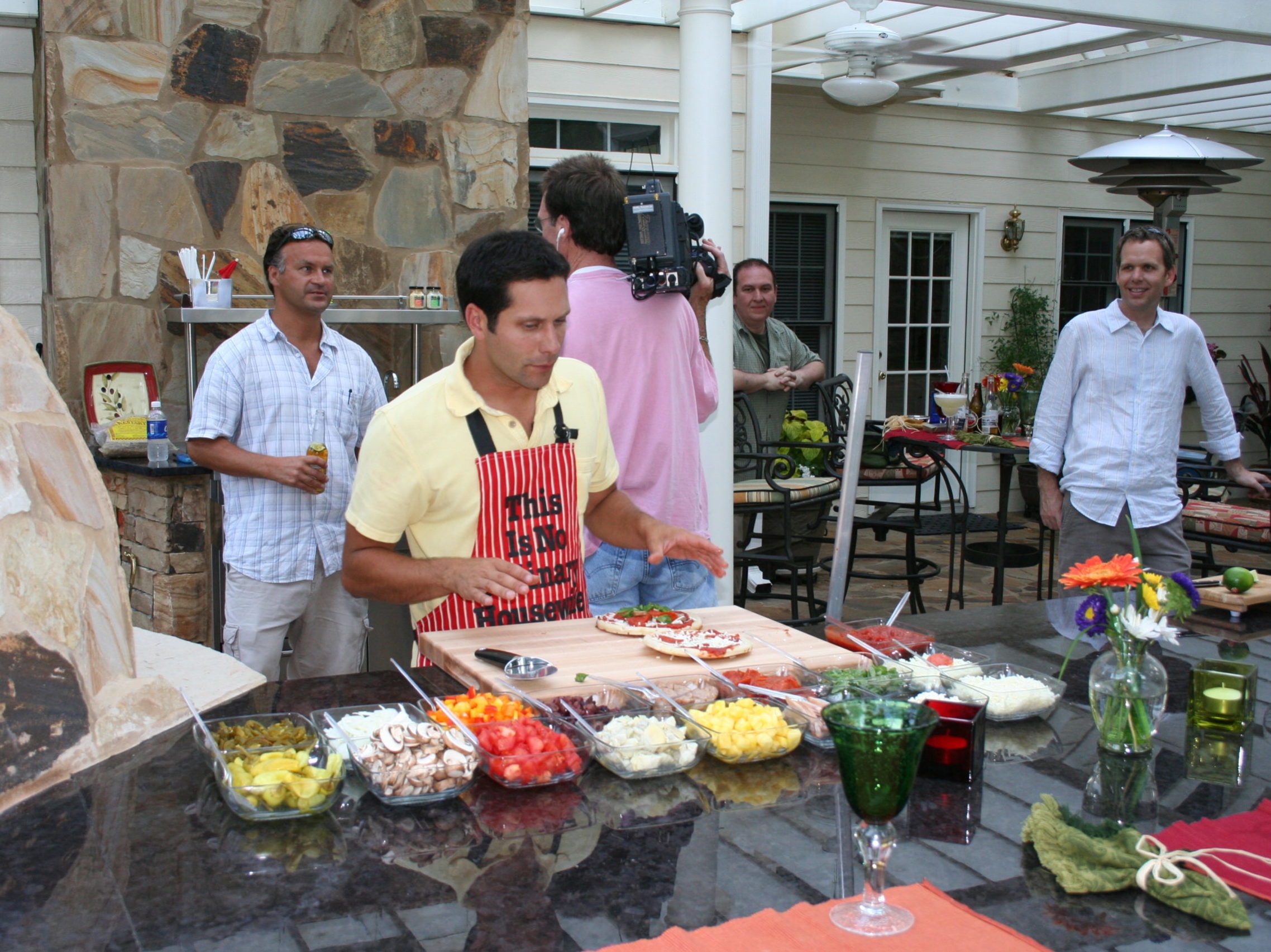 A person in a patterned apron prepares food at an outdoor kitchen counter. Nearby, several others gather, some with a camera, around the patio's stone wall and plants, enjoying the ambiance enhanced by a cozy fireplace.