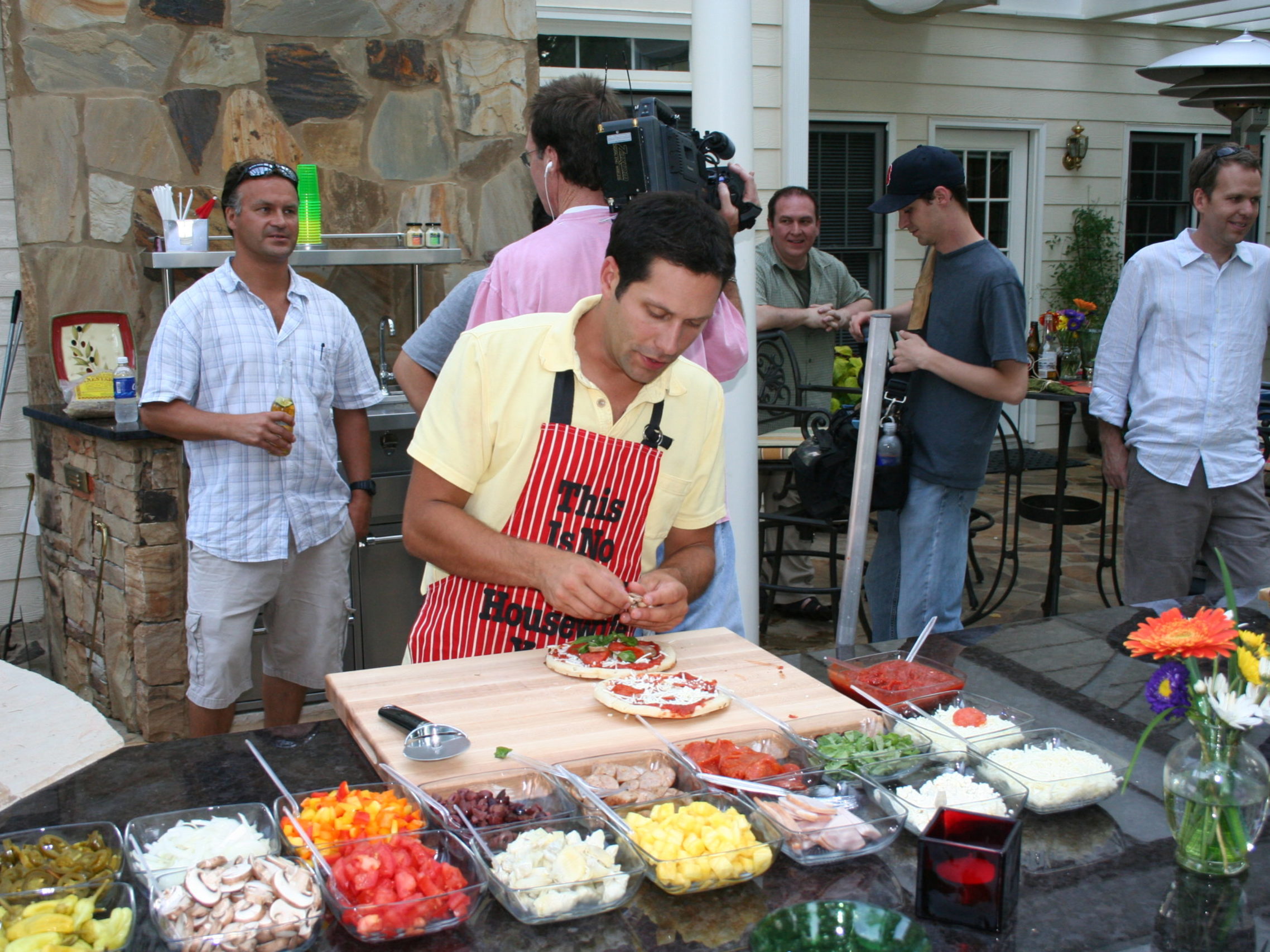 A man in a yellow shirt and apron prepares food at an outdoor kitchen, the warm glow from the nearby fireplace adding charm. Surrounded by fresh ingredients, he captivates several onlookers gathered in the background.