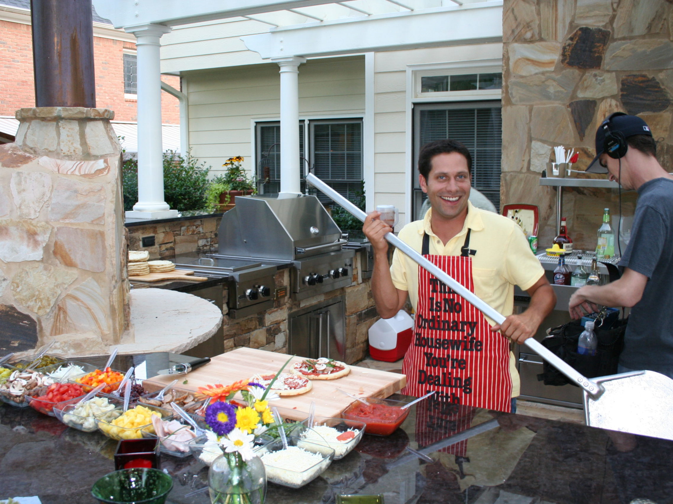 Two men are in an outdoor kitchen beside a mesmerizing stone fireplace. One is holding a pizza peel near the oven, smiling, while the other prepares food. A variety of pizza toppings are spread across the counter, ready to create a delicious masterpiece.