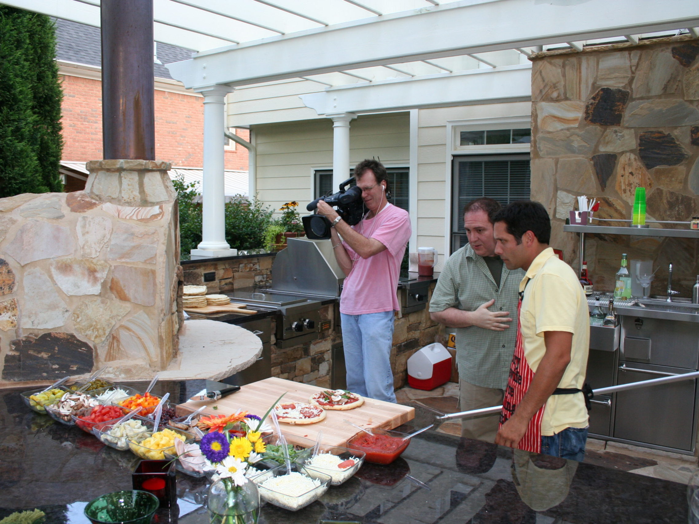 Two men prepare pizza in an outdoor kitchen, their actions carefully captured by another man filming nearby. A variety of toppings and flowers adorn the counter, with the warmth of a fireplace adding a cozy charm to the lively culinary scene.