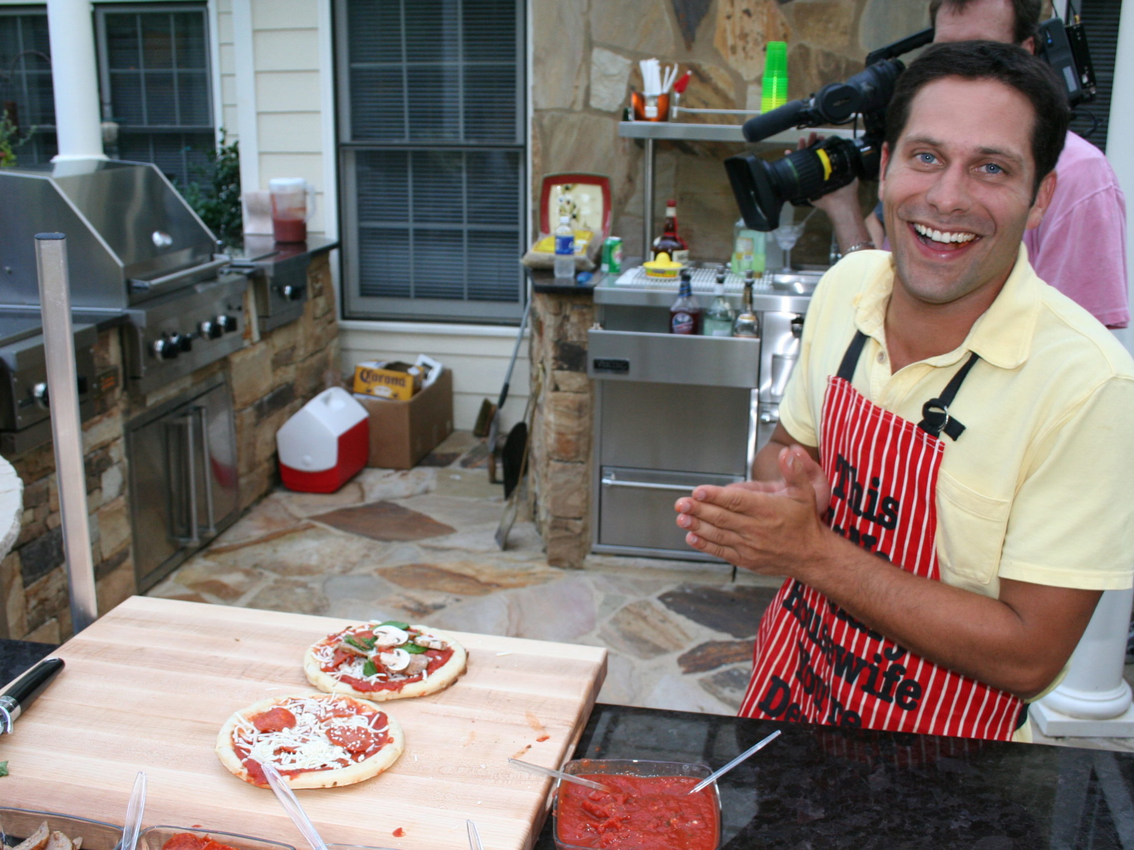 A person in a yellow shirt and red apron smiles beside a kitchen counter with two pizzas, set against the backdrop of an outdoor kitchen and fireplace, surrounded by a barbecue setup and filming equipment.