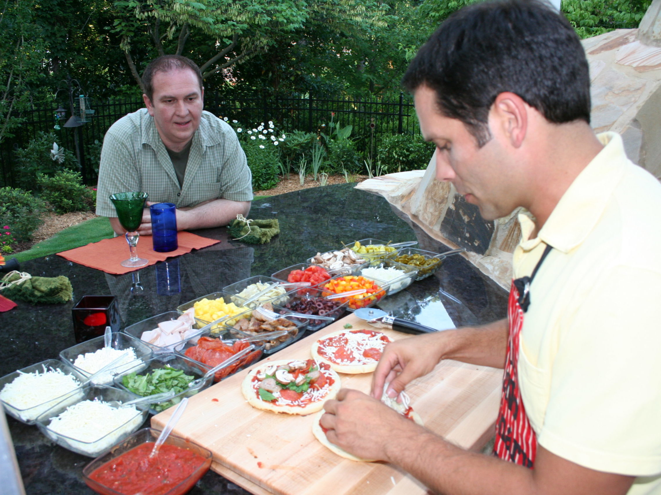 Two men at an outdoor table with assorted pizza toppings. One man is skillfully making a pizza, while the other watches beside a cozy outdoor kitchen. A garden and a charming fireplace are visible in the background, completing this serene culinary setting.