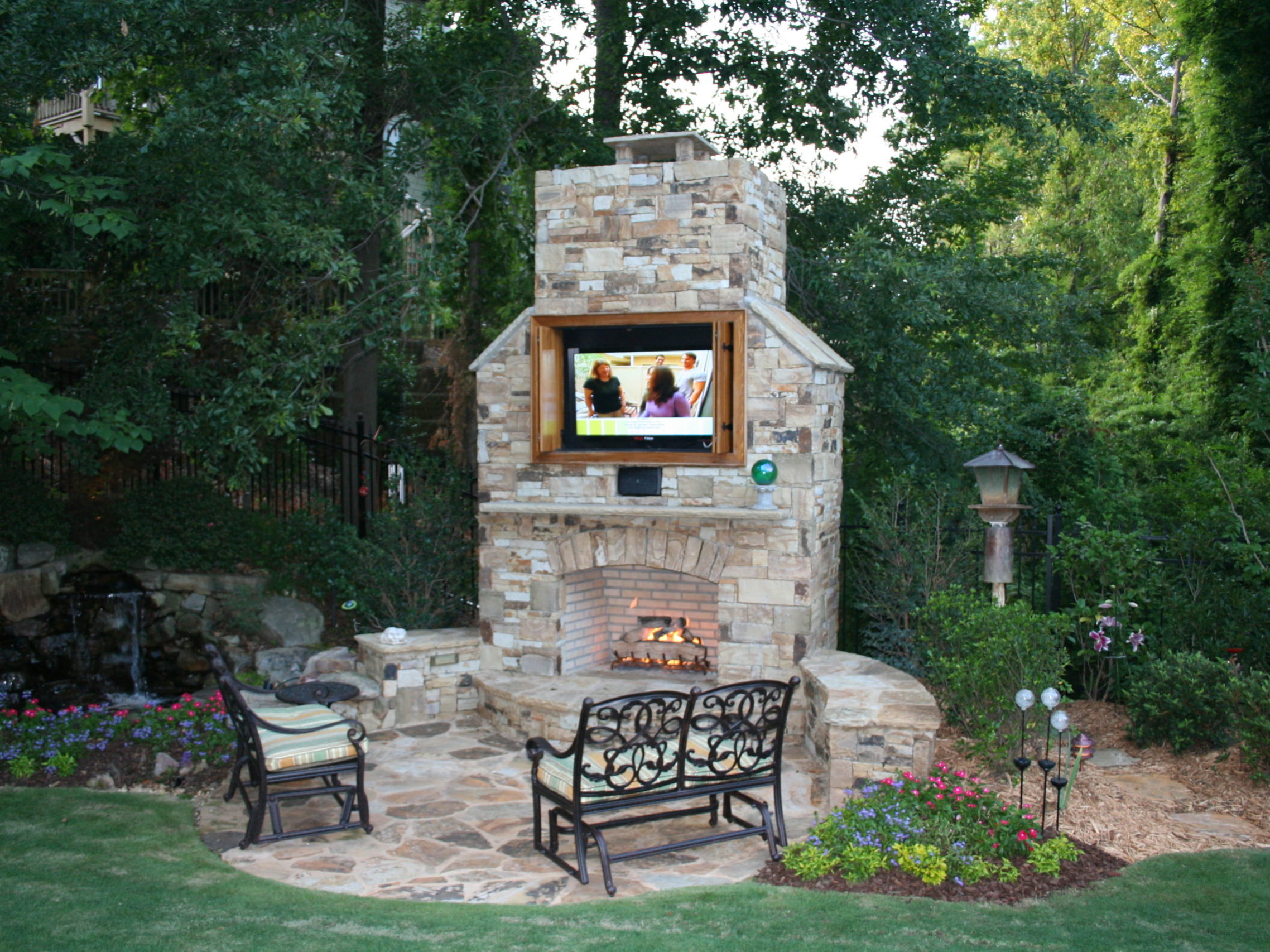 An outdoor kitchen complements the stone fireplace with a mounted TV above it, creating a perfect space surrounded by benches and a landscaped garden area.